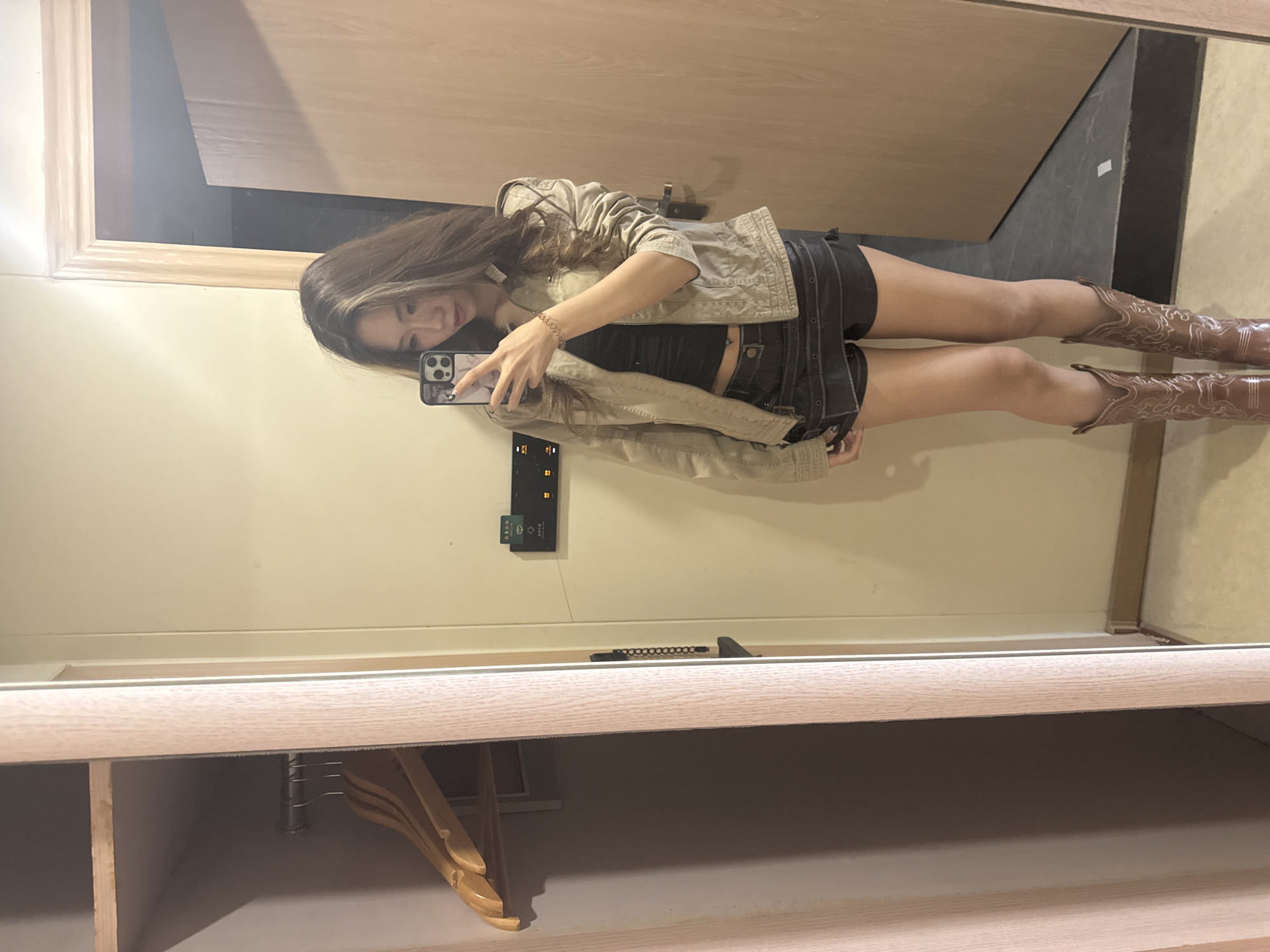A young woman is taking a mirror selfie in what appears to be a hotel room. She is wearing a tan jacket, black top, black shorts, and brown cowboy boots. The background reveals a wooden door, a wall with electrical outlets, and a clothing rack with hangers. The location is identified only by the mention of Dunhuang City, China, which is presumed as there is no further context in the image itself.