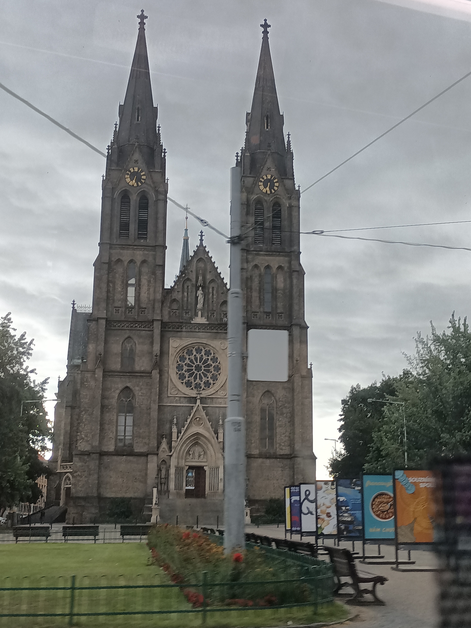 The image shows the Basilica of St. Peter and St. Paul in Vyšehrad, Prague, Czechia. The basilica is a prominent Gothic structure with two tall spires, one of which features a clock face. The sky is overcast, with visible power lines in the foreground. To the right of the basilica, there are several outdoor displays or posters. In front of the basilica, there is a grassy area with benches and flowerbeds. The overall impression is of a city park adjacent to a historical religious building.