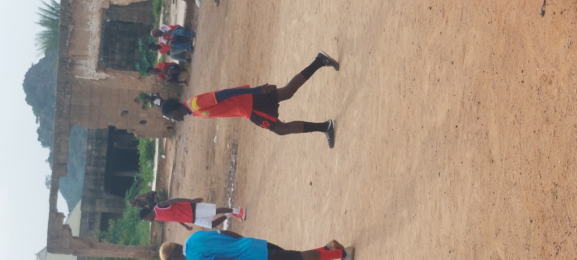 In Bukuru, Nigeria, a young Black male, dressed in a red and yellow soccer jersey, black shorts with red trim, black socks, and black sneakers, is captured in a full-body shot from the knees up. He is in motion, with his left leg bent and his right leg extended backward, suggesting he is walking or jogging. He is positioned in the foreground, facing left in profile, and appears to be in a dirt field. Behind him, a few other individuals are visible. To his left, another young Black male in a bright blue t-shirt is also in motion, facing away from the camera. Further back, a young Black male in a red sleeveless jersey and white shorts is visible, with several other individuals sitting or standing in the background, some wearing red and white shirts. The environment includes a partially ruined brick building with an open archway and a hill with sparse vegetation in the distance. The ground is composed of sandy dirt, with some scattered debris. The sky is overcast, suggesting a cloudy day.