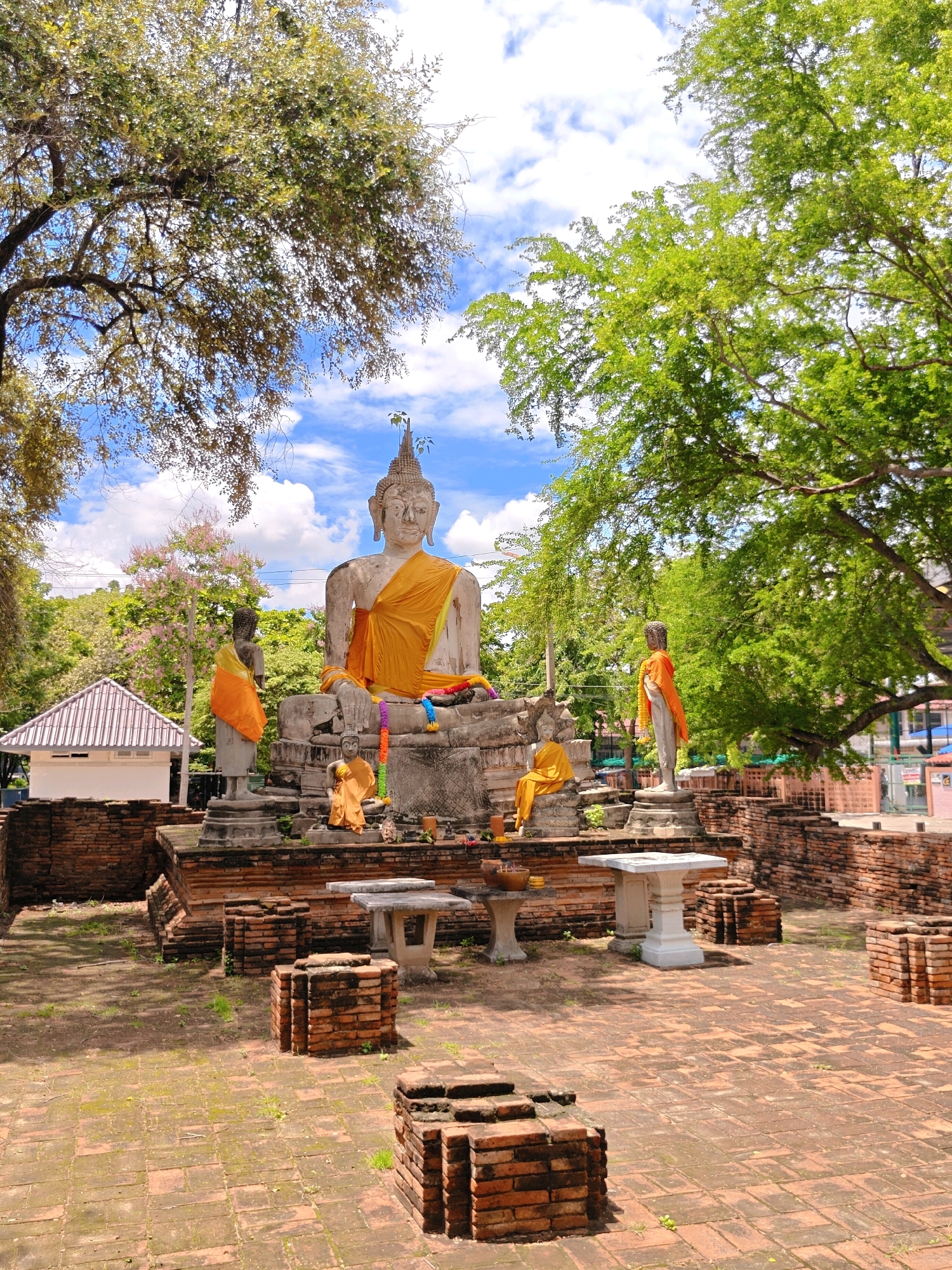 The image depicts a religious site featuring several Buddha statues within a courtyard. The main subject is a large, seated Buddha statue draped in orange cloth. To the left of the large statue, a smaller standing Buddha statue, also draped in orange, is visible. In front of the large Buddha, a small, seated Buddha statue is present. The statues are situated on a raised brick platform, surrounded by a low brick wall. Several small tables or altars are placed within the courtyard. The ground is paved with reddish bricks, interspersed with patches of grass. Lush green trees with dense foliage frame the scene, and a blue sky with scattered white clouds is visible in the background. A small white building with a tiled roof is also present on the left side of the image. The overall setting suggests a temple or historical religious complex. The location is identified as Phra Nakhon Si Ayutthaya City Municipality, Thailand.