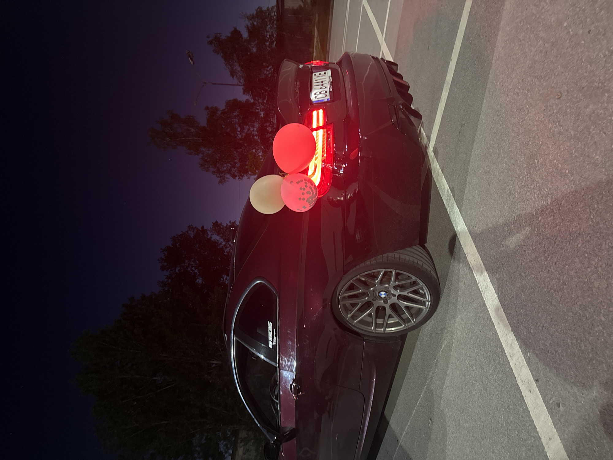 A low-angle, close-up shot depicts the rear passenger-side of a dark-colored BMW sedan parked on asphalt within a parking lot setting in Unknown, Unknown. The vehicle is adorned with three balloons of varying colors—red, white, and pink with a speckled pattern. The vehicle's taillights are illuminated, and the license plate, "CB 844WE," is visible. The car's rear tire features a distinctive BMW wheel design. The parking lot is marked with painted white lines. In the backdrop, trees are silhouetted against a dark blue night sky with a street light visible in the distance.