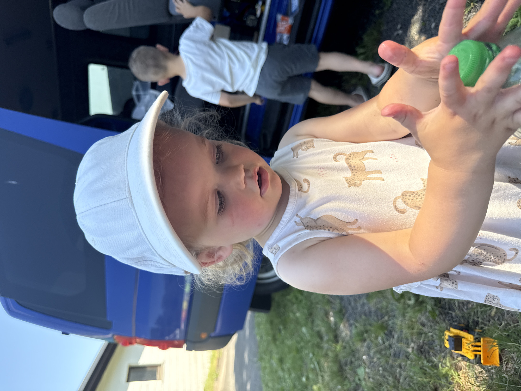 A young child wearing a white baseball cap and a white sleeveless dress with a leopard print is in the foreground, looking down at their hands which are holding a green-capped bottle. To the left, a yellow toy construction vehicle is on the grass. In the background, a blue van is parked, and another child wearing a white t-shirt and grey shorts stands near the open rear door of the van. The setting appears to be an outdoor area, possibly a residential yard, with grass and a paved surface. The lighting suggests it is daytime, with sunlight casting shadows. Beltershain, Germany is specified as the location.