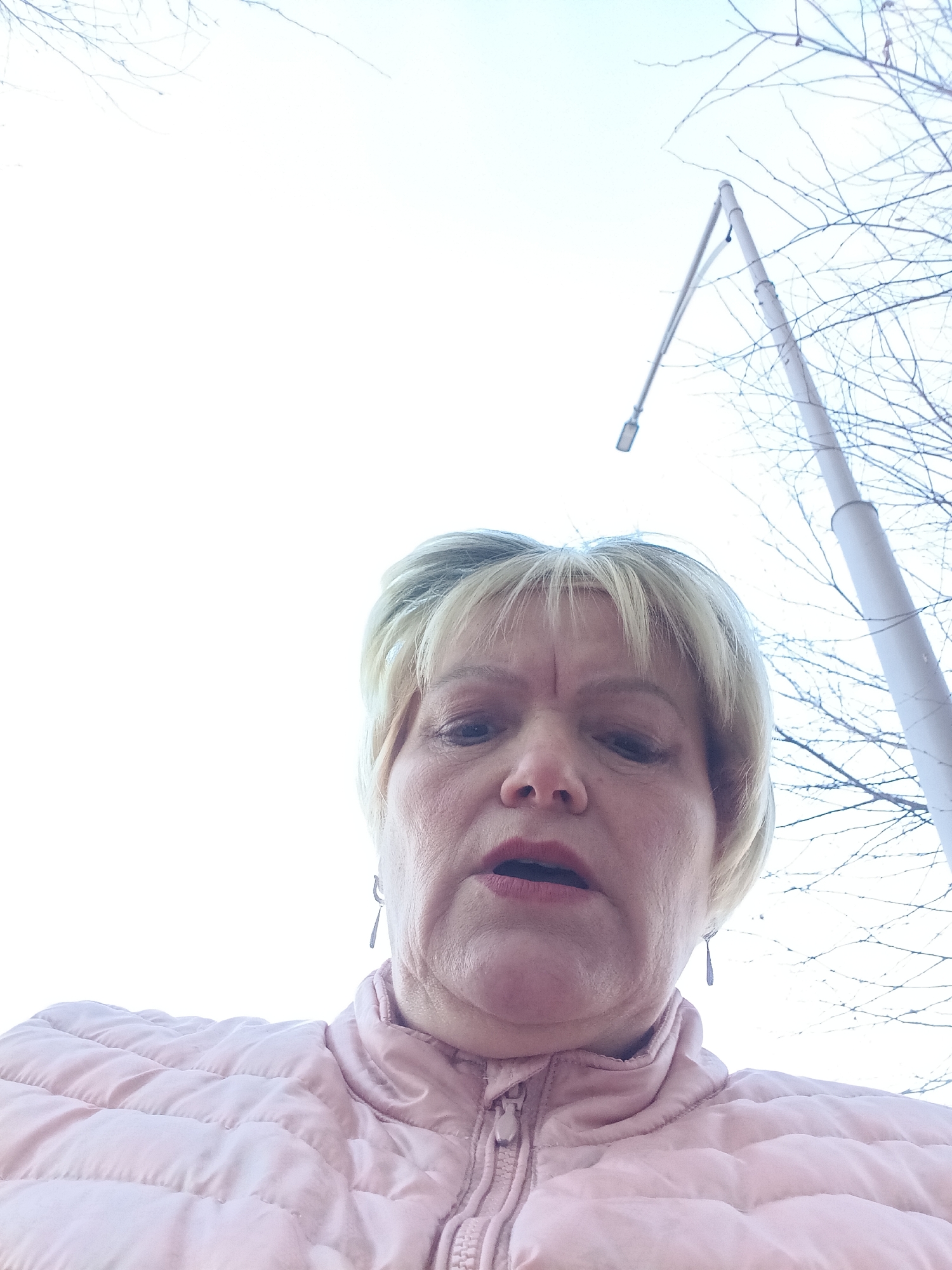 A low-angle outdoor portrait depicts a middle-aged blonde woman with short hair and bangs, looking downwards towards the camera. She is wearing a light pink, possibly quilted or puffer-style, zippered jacket. A silver-colored, teardrop-shaped earring is visible on her right ear. Her mouth is slightly open. The background consists of a bright, overcast sky. To the right, a tall, light-colored street lamp post with a horizontal arm and a rectangular light fixture extends into the frame. Bare tree branches are visible against the sky, primarily in the upper right quadrant. The scene is located in Odintsovo, Russia.