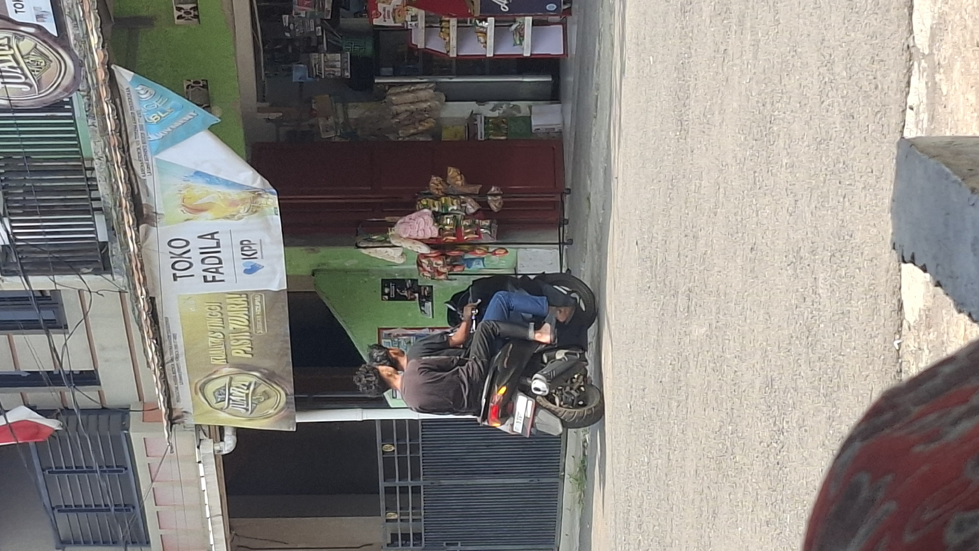 The image shows two individuals riding a black scooter on a paved road in front of a building in Cilaku, Indonesia. The building has a green facade and a banner displaying "TOKO FADILA" and "JUARA". The individuals are wearing casual clothing, with one person in a black t-shirt and the other in a black long-sleeved shirt. Both are facing forward and appear to be riding the scooter. Behind them, there are various items displayed on racks outside the store, suggesting it is a small shop. The environment appears to be a typical street scene in a town or city.