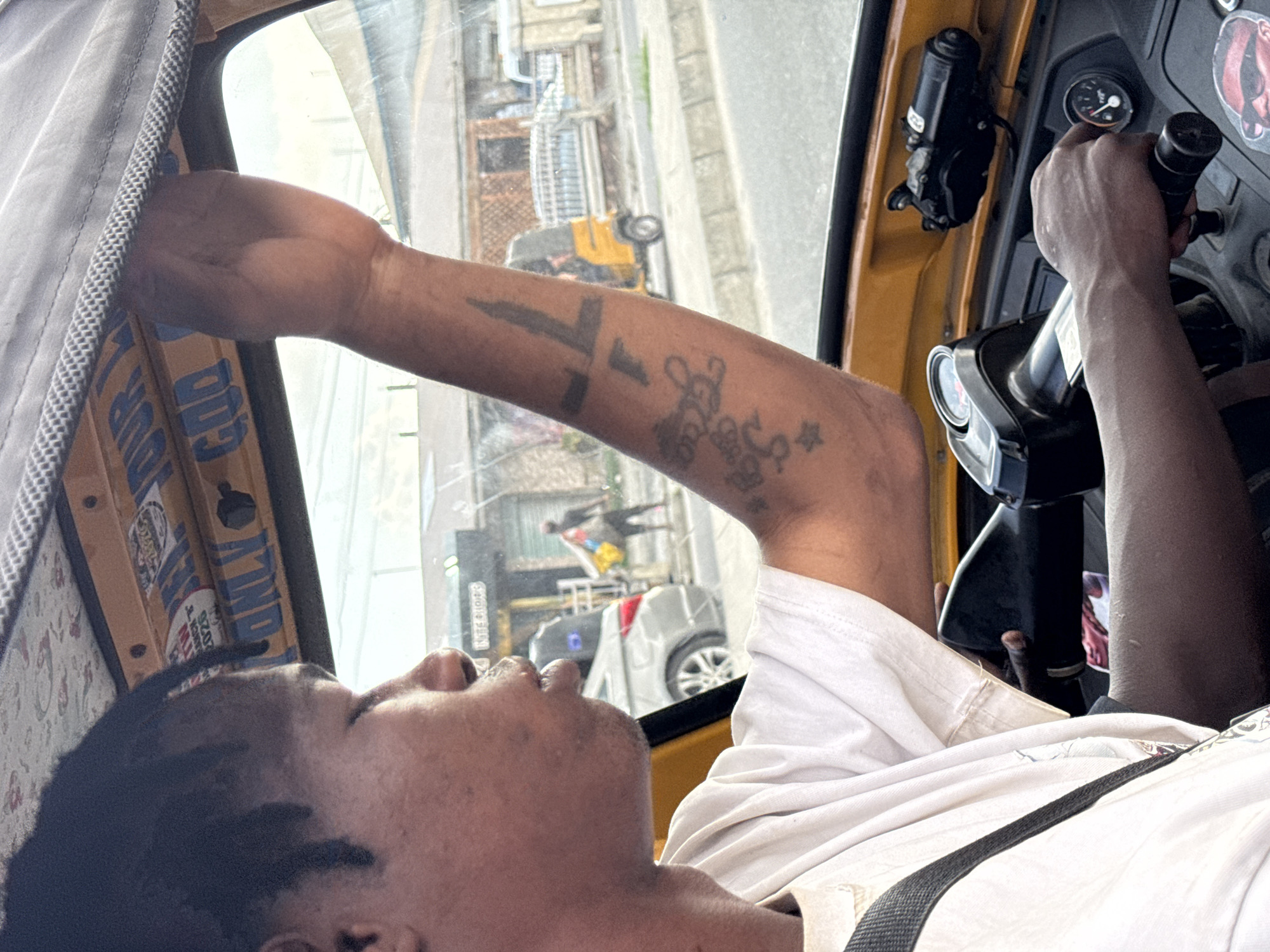 A medium, eye-level shot from inside a commercial tricycle, or "keke," in Ado, Nigeria. The driver, a dark-skinned man with braided hair, is seen from the chest up. He is wearing a white t-shirt with a black strap across his chest. His right arm is extended, gripping the roof of the vehicle, while his left hand operates the steering mechanism. Tattoos are visible on his right forearm. The tricycle's interior is yellow, with stickers on the window frame. Outside, the street scene reflects in the window, showing other vehicles, pedestrians, and buildings. A yellow tricycle is visible further down the road.