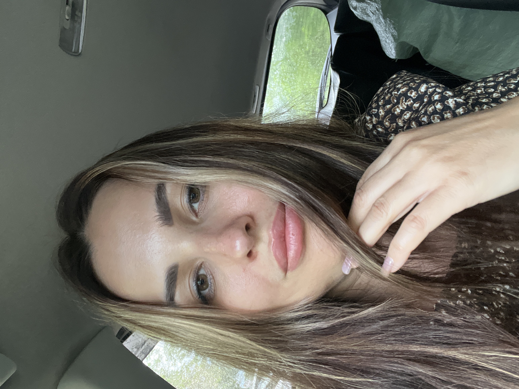 A medium close-up shot shows a woman with long, wavy brown hair with blonde highlights, with her face turned slightly towards the camera. She is wearing a dark top with a subtle print. Her left hand, with light pink nail polish, is raised and touching her hair near her face. The background appears to be the interior of a car, with a window showing a blurry green outdoor scene, suggesting it might be raining. The lighting is natural and diffused.