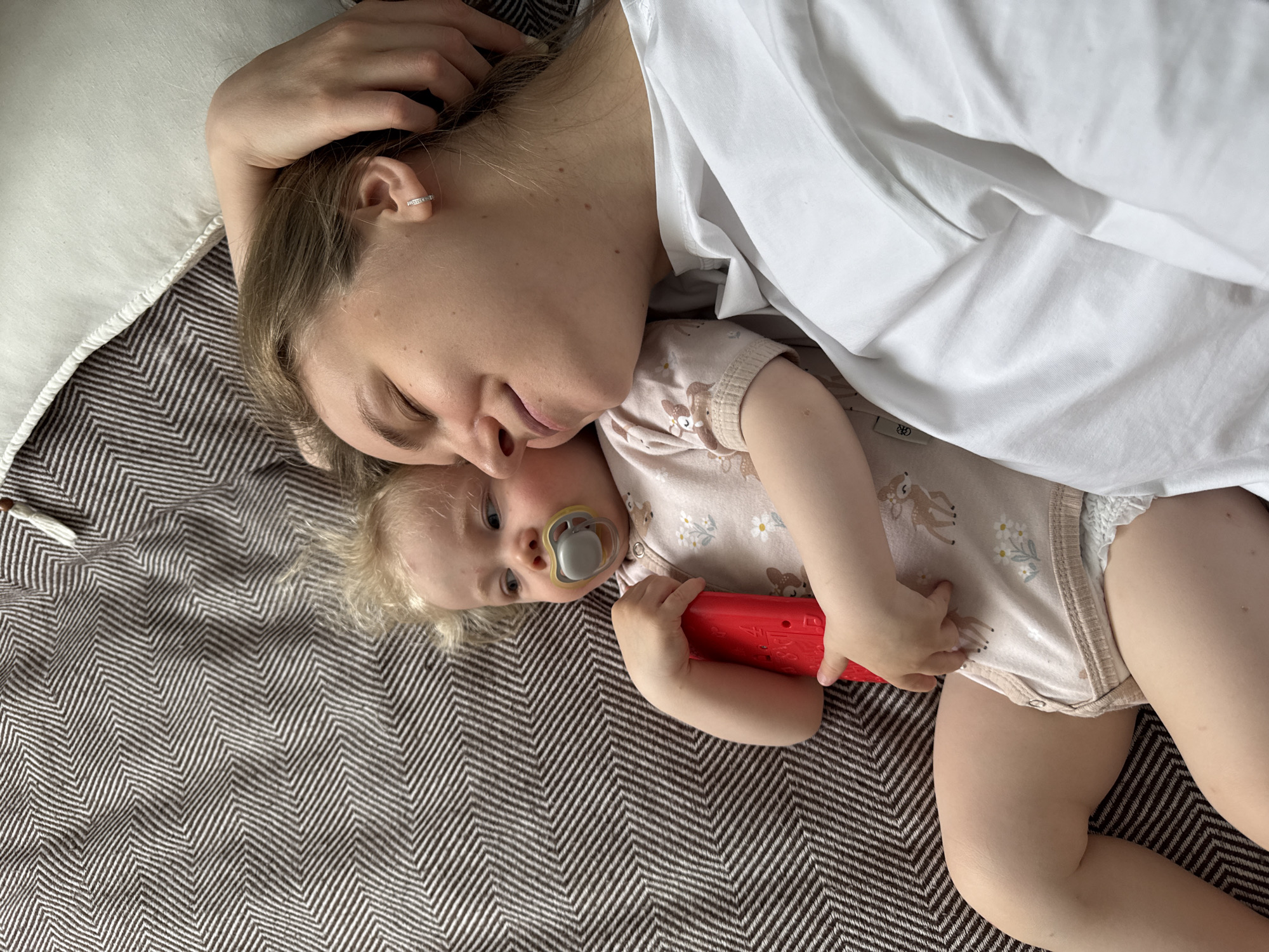 A fair-skinned woman lies on her side on a brown and white herringbone-patterned surface, a white pillow behind her head, and covered partially by a white fabric. Adjacent to her, a fair-skinned baby, also lying down, clutches a red object. The baby has a pacifier in their mouth. The overall environment suggests a casual, indoor setting. The location of the city of Vladimir, Russia is not indicated in the image.