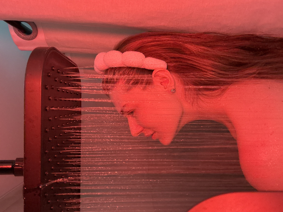A woman with auburn hair is shown in profile, standing under a modern, multi-spray shower head. She wears a white, plush headband. Water streams directly into her face as she lowers her head, eyes closed. The scene is bathed in a deep red light, giving the image an intense and warm tone. The woman's skin appears damp and flushed, highlighting the direct contact with the shower's spray.