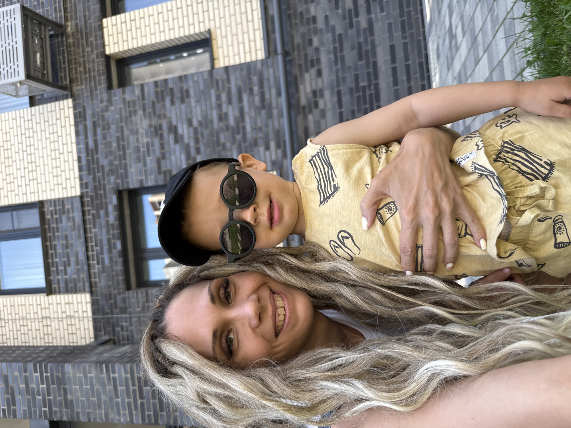 A woman with blonde dreadlocks and a young child wearing sunglasses and a cap are posing for a selfie outdoors in front of a modern brick building. The woman is smiling at the camera, her arm around the child. The child is looking away from the camera. The building has dark and light brickwork and windows. The ground in front of the building is paved with light-colored tiles and a strip of grass.