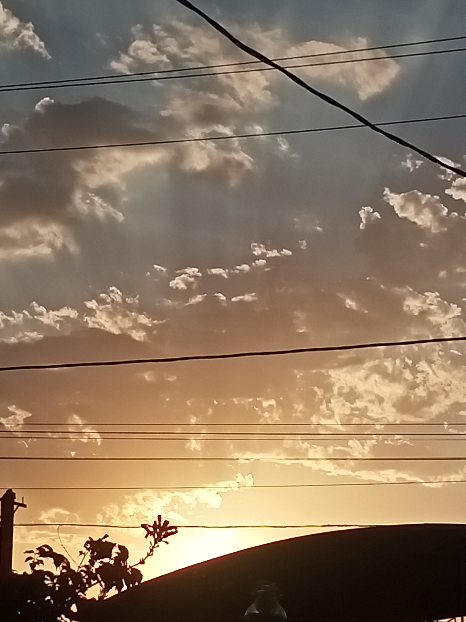 The image shows a silhouetted scene against a sunset sky, with power lines crossing the frame. In the foreground, the dark silhouettes of trees and the edge of a dark roof are visible. A utility pole is also silhouetted on the left. The sky is filled with scattered clouds, illuminated by the warm hues of the setting sun. The colors range from pale blue at the top to orange and yellow towards the horizon. The scene appears to be an outdoor view within the city of Sokuluk, Kyrgyzstan.