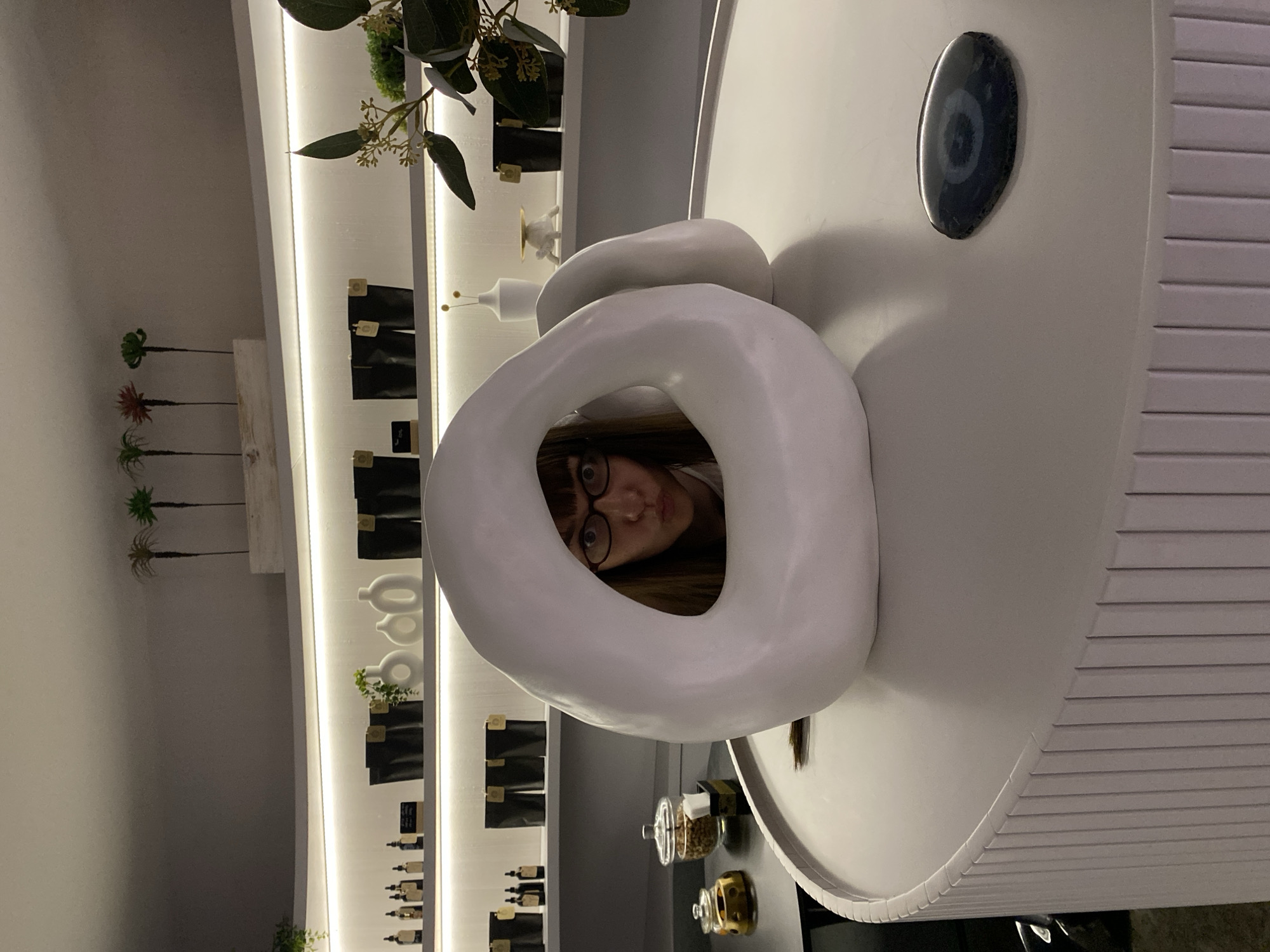 A woman wearing glasses is peering through a white donut-shaped object. The scene appears to be indoors, possibly in a store or gallery. There is a counter with various items, including jars, behind the donut object. To the left, a wall displays shelves with packaged products and decorative plants. The provided location, Votkinsk, Russia, is not apparent within the visible content of the image.