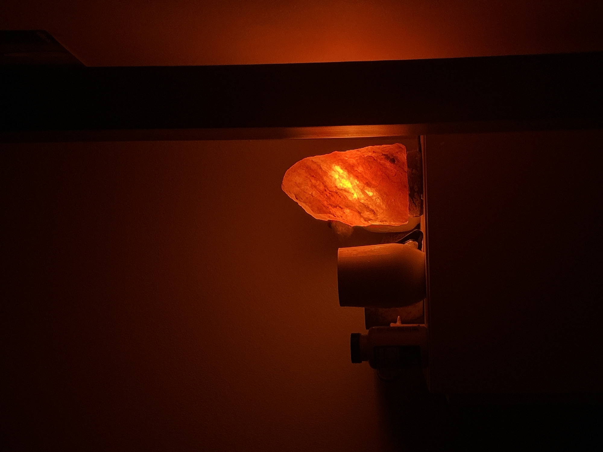 A Himalayan salt lamp is mounted on a wall in a dimly lit room. The lamp's rough, crystalline surface glows with a warm orange light, illuminating the textured walls around it. The light emanates from within the rock salt, creating a soft, ambient glow. The lamp appears to be a decorative item, adding a warm and relaxing atmosphere to the space. The wall itself is a muted orange color, blending with the lamp's light. In the background, a darker area suggests a shadow or a recess in the wall. The overall scene conveys a sense of tranquility and warmth. The image was likely taken indoors, possibly in the evening or in a room with limited natural light, given the prominent use of the lamp for illumination.