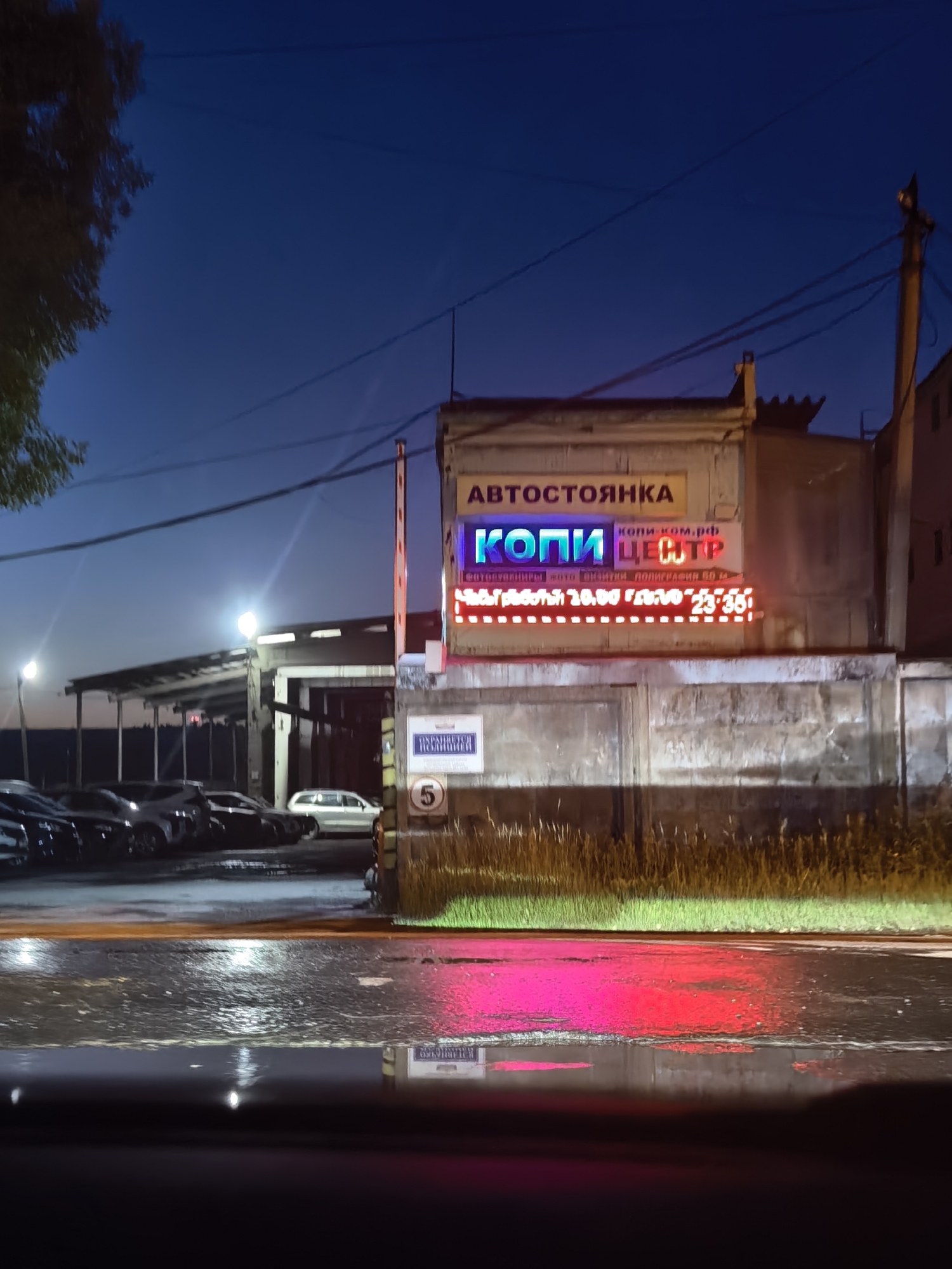 The image depicts a neon sign for "КОПИ ЦЕНТР" (Copy Center) on a building at dusk or night. The sign is illuminated with bright blue and pink neon lights. Above it, a yellow sign reads "АВТОСТОЯНКА" (Parking Lot). Below the neon sign, a red LED display shows operating hours: "Часы работы 10:00 23:30". A sign on the wall beneath the LED display reads "ОПЛАТИТЬ СТ-ЦИИ" (Pay for parking). The scene appears to be an outdoor parking lot with several cars parked under a covered area to the left. The ground is wet, reflecting the neon lights. In the foreground, a portion of a car's interior is visible, suggesting the photo was taken from within a vehicle. The sky is dark blue.