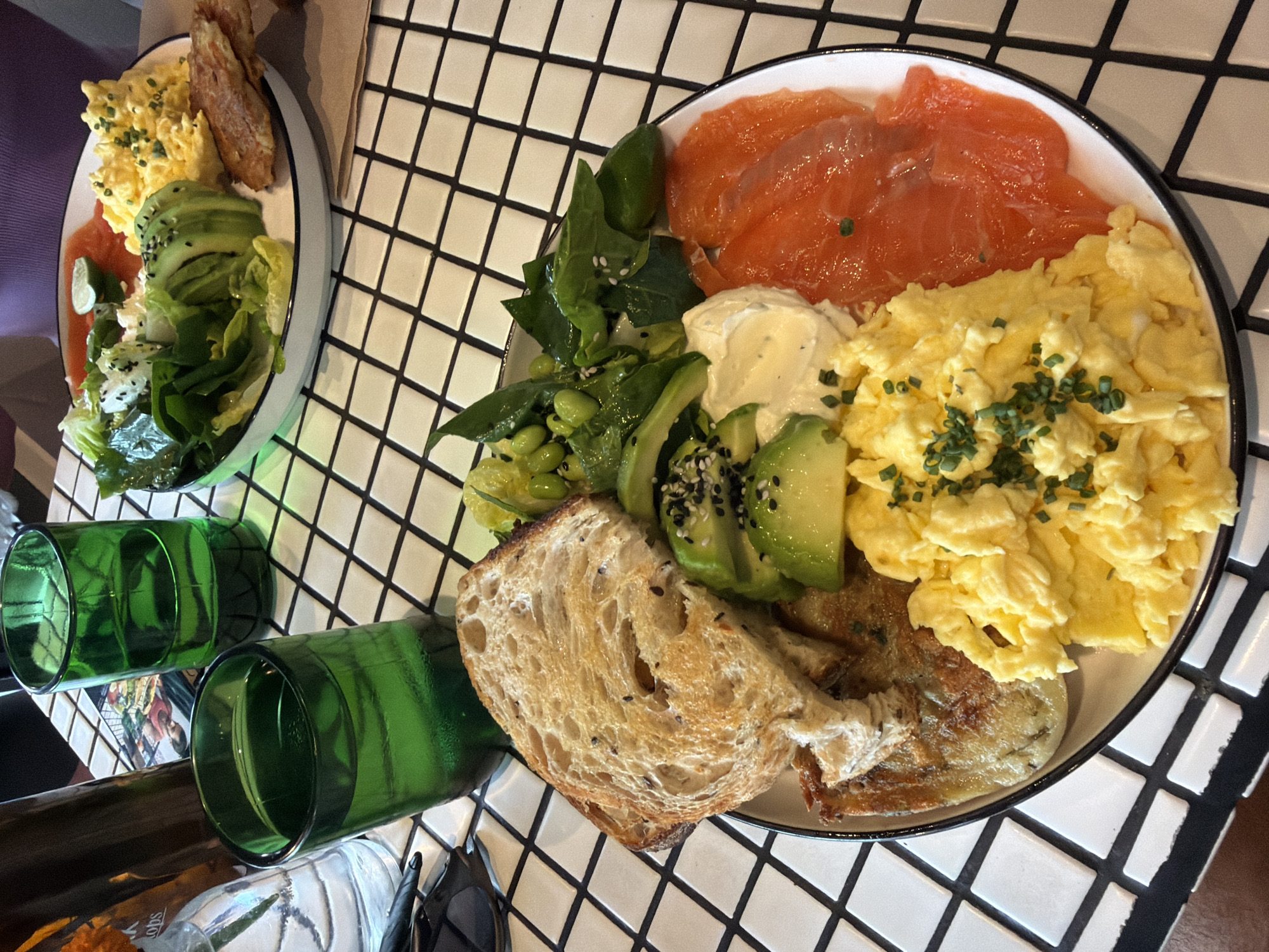 A close-up overhead shot captures a delectable brunch spread on a black and white checkered table. The main dish, a circular plate, is filled with an assortment of savory items. Fluffy scrambled eggs, generously sprinkled with chopped chives, occupy the lower right quadrant. Adjacent to the eggs are vibrant slices of smoked salmon, glistening under the light. To the left of the salmon and eggs, a dollop of creamy white cheese is nestled beside a portion of sliced avocado, which is seasoned with black sesame seeds. A slice of toasted sourdough bread, with a slightly charred crust and visible seeds, rests on top of a golden-brown potato hash. In the upper portion of the plate, a small pile of green leaves, edamame, and possibly cucumber slices creates a fresh salad element.
In the background, a second, smaller white plate holds a similar arrangement of salad, avocado, scrambled eggs, and smoked salmon, suggesting another diner or a larger brunch order. Two dark green glasses, one half-filled with a clear liquid and the other mostly full, stand on the checkered surface to the left. The lighting is bright, illuminating the food and creating a cheerful atmosphere. The setting appears to be a restaurant or cafe, with the checkered tablecloth being a prominent decorative element. The overall scene evokes a sense of a leisurely and satisfying meal.