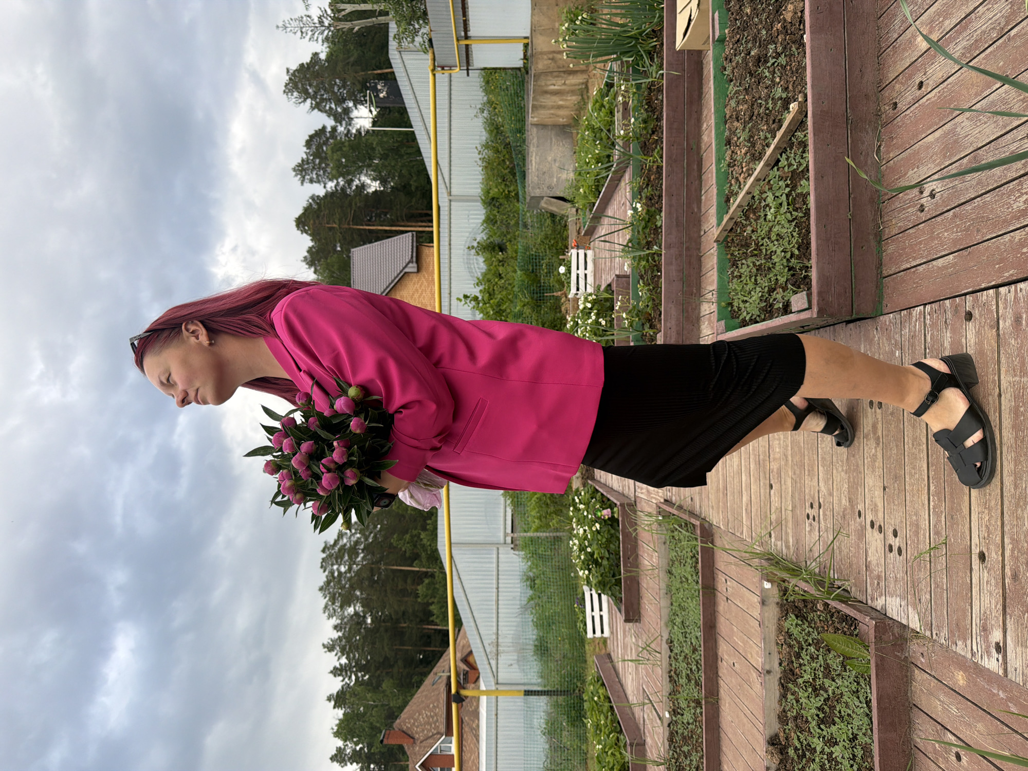 A woman stands on a wooden walkway adjacent to raised garden beds in Chaikovsky, Russia, under a cloudy sky. She is wearing a pink blazer, black dress, and black sandals. She holds a bouquet of pink flowers. A fence runs along the back of the scene, behind which are trees and houses.