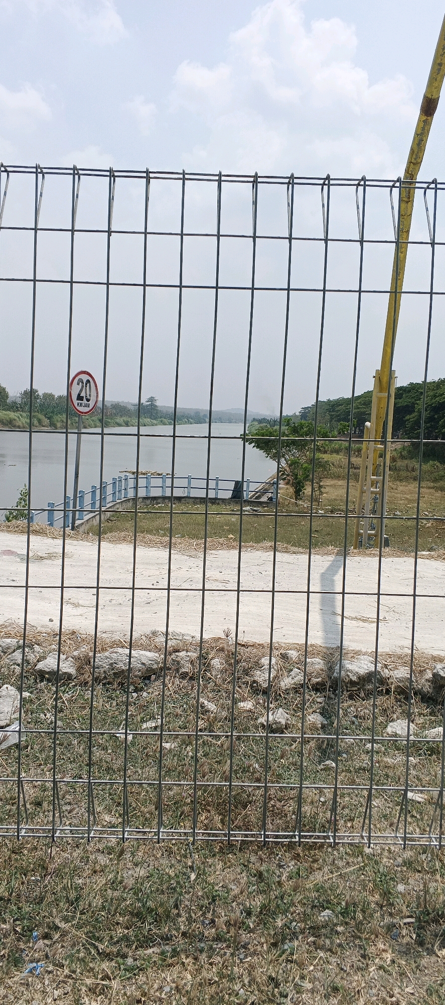 A wire fence is in the foreground of a landscape image. Through the fence, a body of water is visible, with a blue railing along its edge. A speed limit sign indicating "20 KM/JAN" is on a pole to the left of the water. In the background, trees and hills are seen under a cloudy sky. The scene is in Parengan, Indonesia.