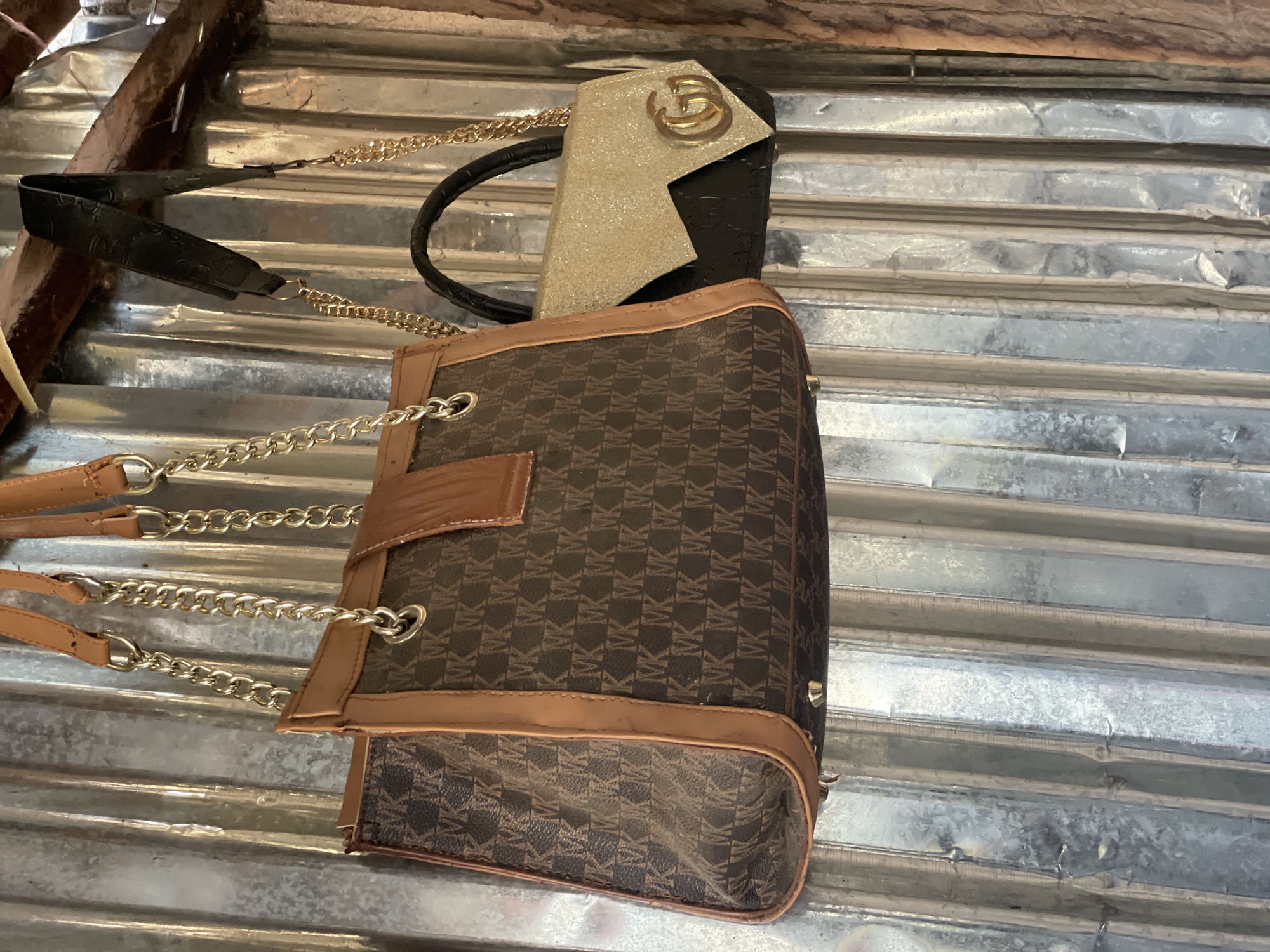 The image shows two handbags hanging on a corrugated metal wall. The handbag on the left is brown with a repeating pattern of "MK" logos and tan leather trim. It has gold chain straps and tan leather shoulder straps. The handbag on the right is black and gold, with a glittery gold front flap and a gold "GG" logo. It also has a gold chain strap. The background is a weathered, corrugated metal surface, likely an exterior wall. The scene appears to be in an outdoor or semi-outdoor setting, possibly a market stall or a storage area in Jalingo, Nigeria.