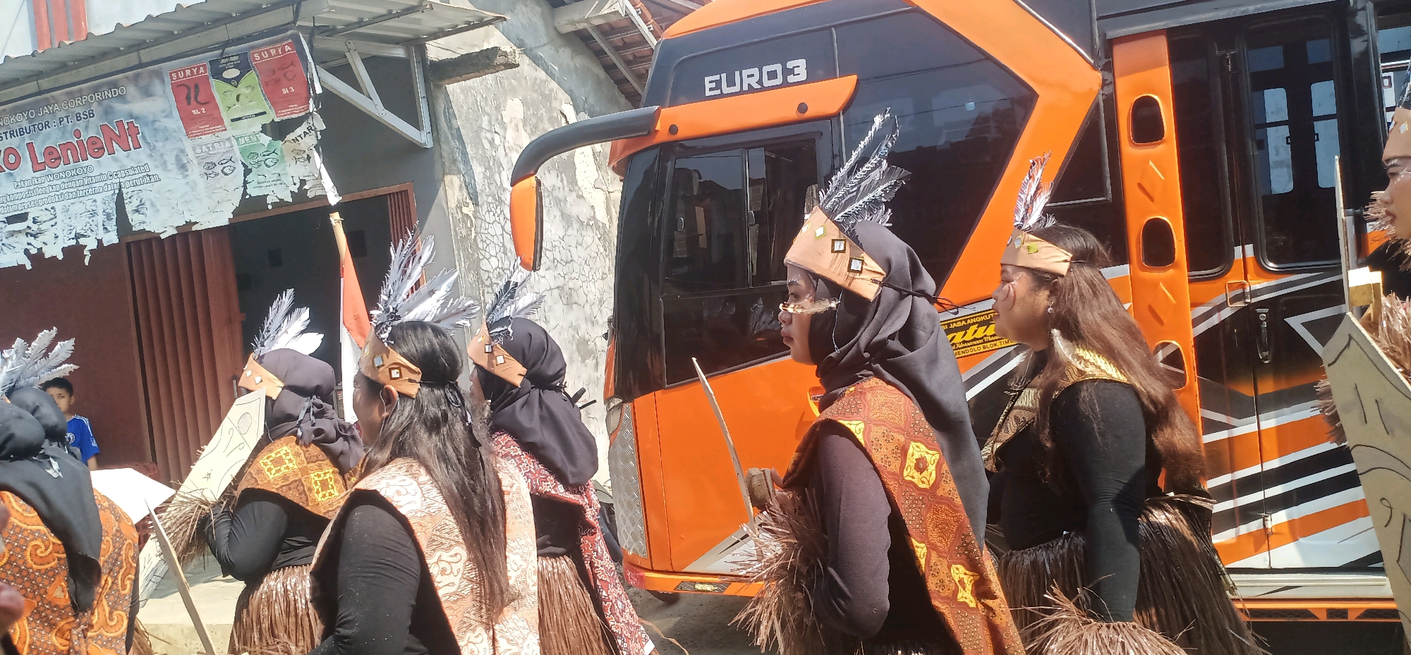 In Kincang, Indonesia, a group of people are participating in a cultural parade. The individuals are adorned in traditional attire, featuring headdresses with feathers, patterned fabrics, and skirts made of woven materials. They are standing in front of an orange and black bus with "EURO 3" written on its side, suggesting a modern transportation element within the traditional event. The background shows a building with a sign that appears to be an advertisement. The overall scene depicts a lively cultural celebration or procession in a street setting.
