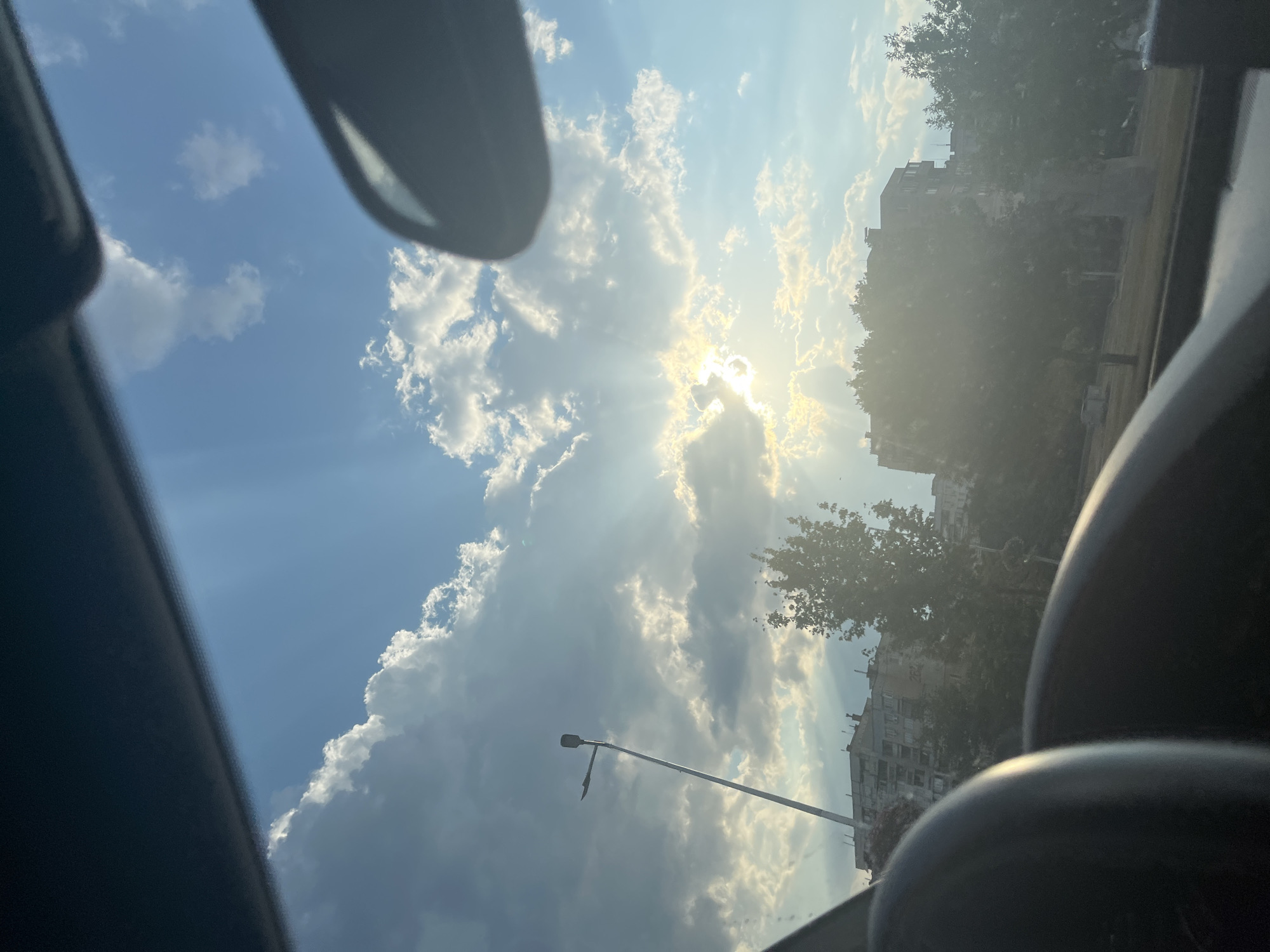 The image captures a view through a vehicle's windshield of a sunny sky with scattered clouds. Sun rays pierce through the clouds, creating a bright, radiant effect. In the mid-ground, a tall street lamp stands to the left, and several apartment buildings and trees are visible. The foreground is dominated by the interior of the car, with parts of the dashboard and possibly a rearview mirror or sun visor visible. The scene appears to be set in an urban environment, likely in the city of Montana, Bulgaria, given the architectural style of the buildings and the general atmosphere. There are no people or specific actions depicted in the image.