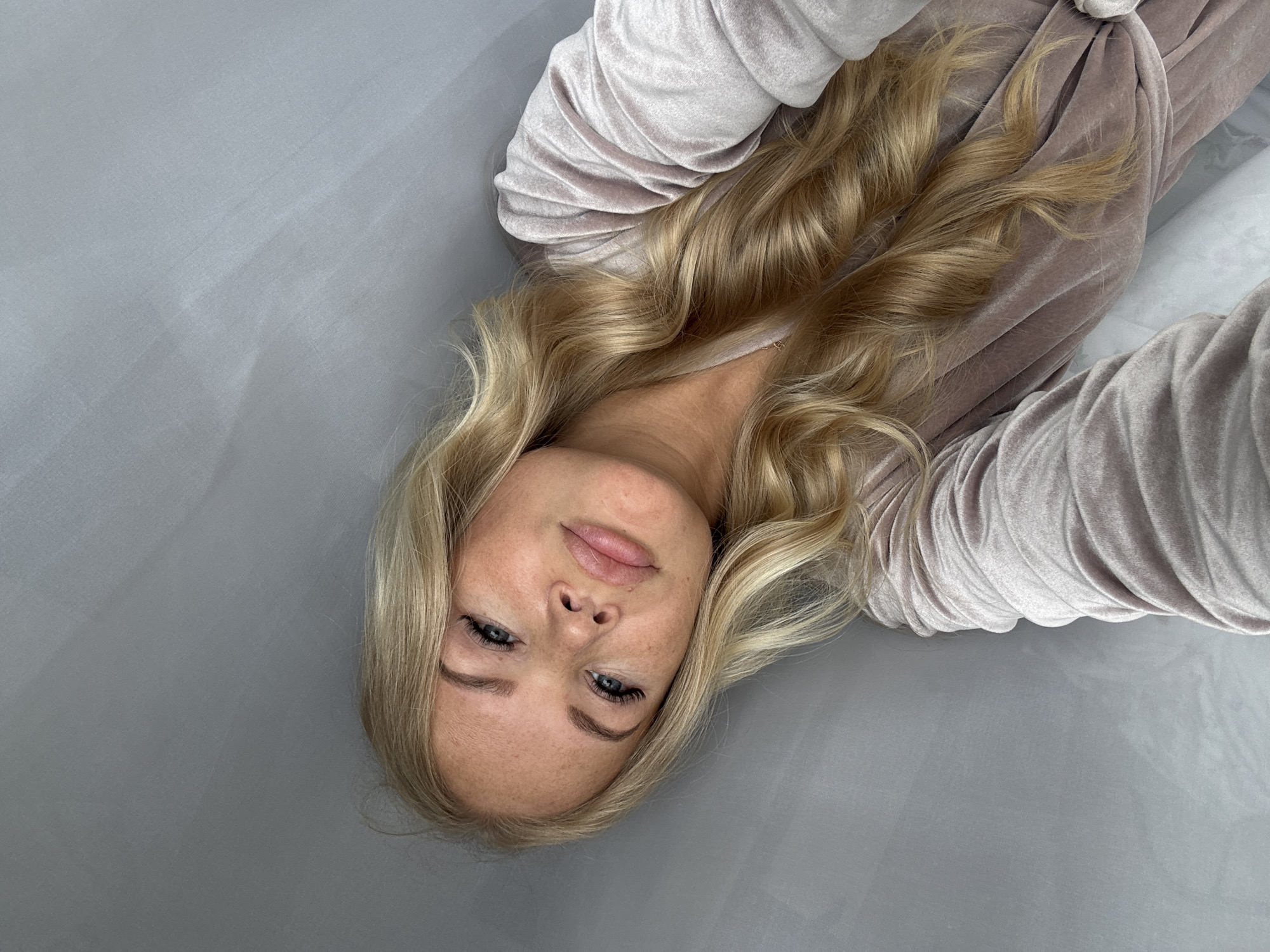A fair-skinned woman with blond, wavy hair is lying on a grey surface, presumably cloth, and is partially dressed in what appears to be a light purple, velvet-like robe. Her face is slightly angled towards the viewer, displaying visible freckles on her forehead and nose. She is wearing light makeup, with a focus on her eyelashes and lips. Her eyes are a light blue or grey color. The composition of the image suggests it was taken from an overhead angle. The location is unspecified.