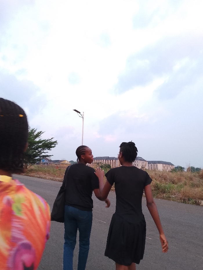 Three women are walking on a paved road in Umueshi, Nigeria. Two women in the center of the image are walking side-by-side. The woman on the left is wearing a black shirt and blue jeans, carrying a black bag on her left shoulder, while the woman on the right is wearing a black dress. A third woman, partially visible on the left edge of the image, is wearing a colorful shirt. In the background, a building is partially obscured by dry vegetation. A street light is also visible. The sky is overcast.
