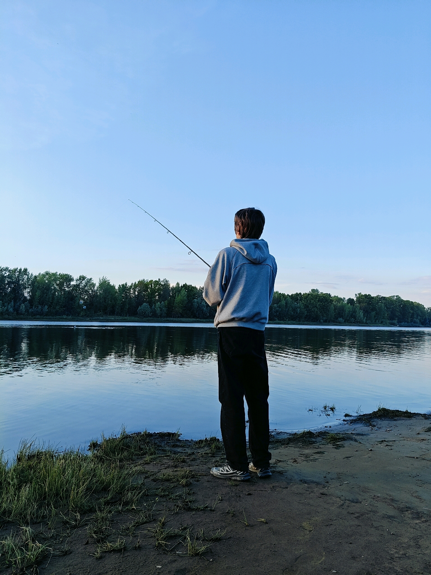 A lone individual stands on the muddy bank of a wide river, facing away from the camera and casting a fishing line. The person is wearing a grey hooded sweatshirt and dark pants. The river is calm, reflecting the clear blue sky and the dense green treeline on the opposite bank. The environment suggests a natural, undeveloped area, possibly a park or a less populated section of the city. The time of day appears to be late afternoon or early evening, given the soft lighting.