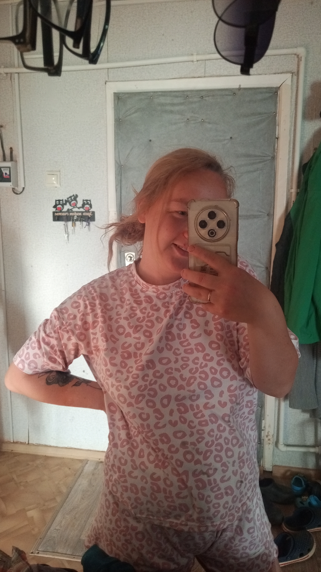 A fair-skinned person with reddish-blonde hair is holding a smartphone to take a selfie in what appears to be an indoor setting. The person is wearing a pink and white leopard-print pajama set, consisting of a short-sleeved t-shirt and shorts. A tattoo is visible on the upper part of their left arm. A wedding ring is visible on the fourth finger of their left hand.
In the background, there is a door with a grayish covering. Hanging from the wall are a key holder shaped like owls and several pairs of glasses. Shoes are scattered on the floor near the bottom of the frame. A green coat and gray garment are visible.
The environment appears utilitarian and domestic, with no clear indication of a city or specific location.