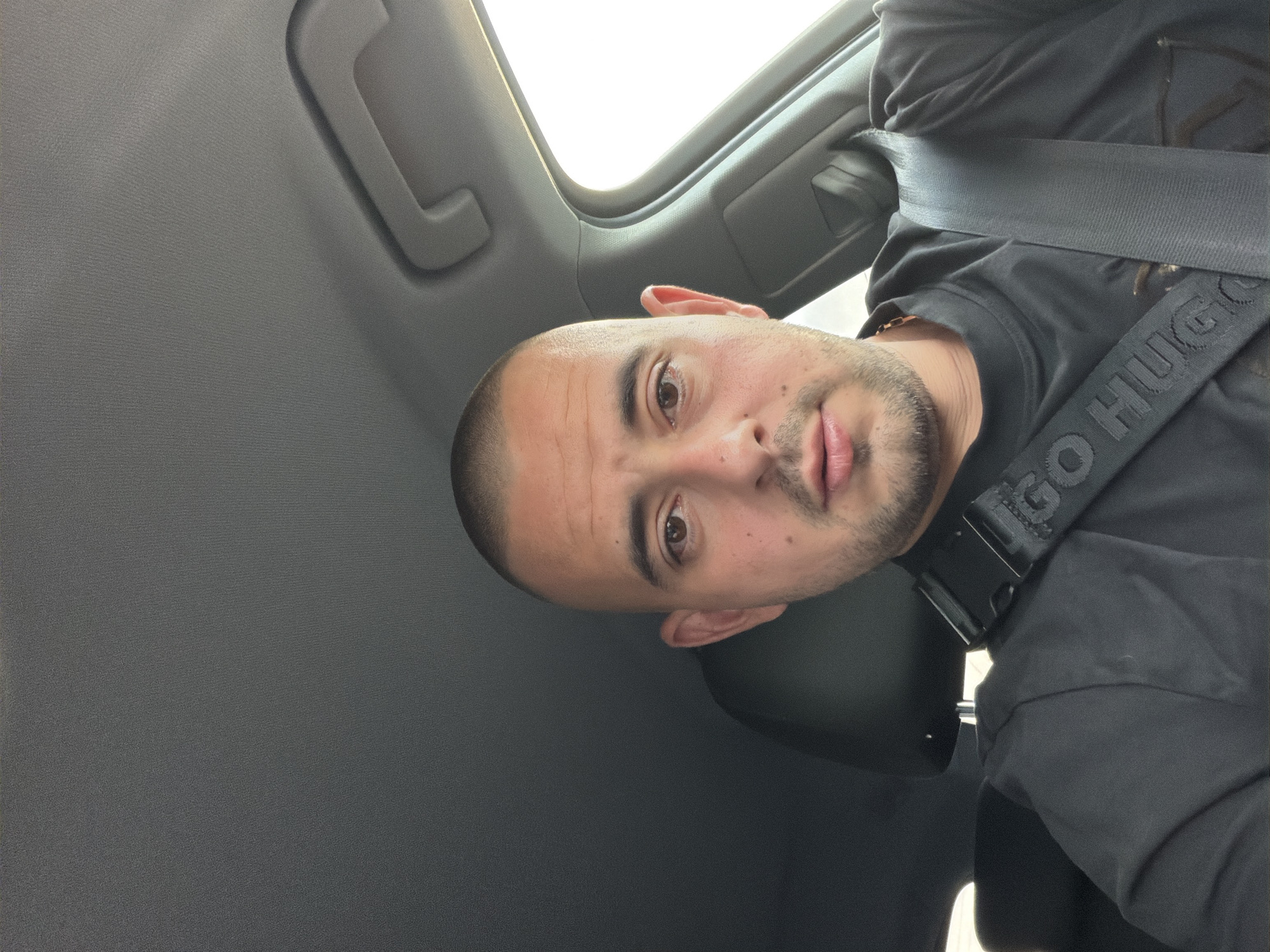 The image is a selfie taken from inside a car. A man with a shaved head and a short beard is in the driver's seat, wearing a black t-shirt and a black seatbelt with "HUGO" written on it. The car interior is visible, with the grey ceiling and a handle on the right side of the frame. The background shows a bright, possibly sunny sky through the car window. The location is not specified in the image or provided context.