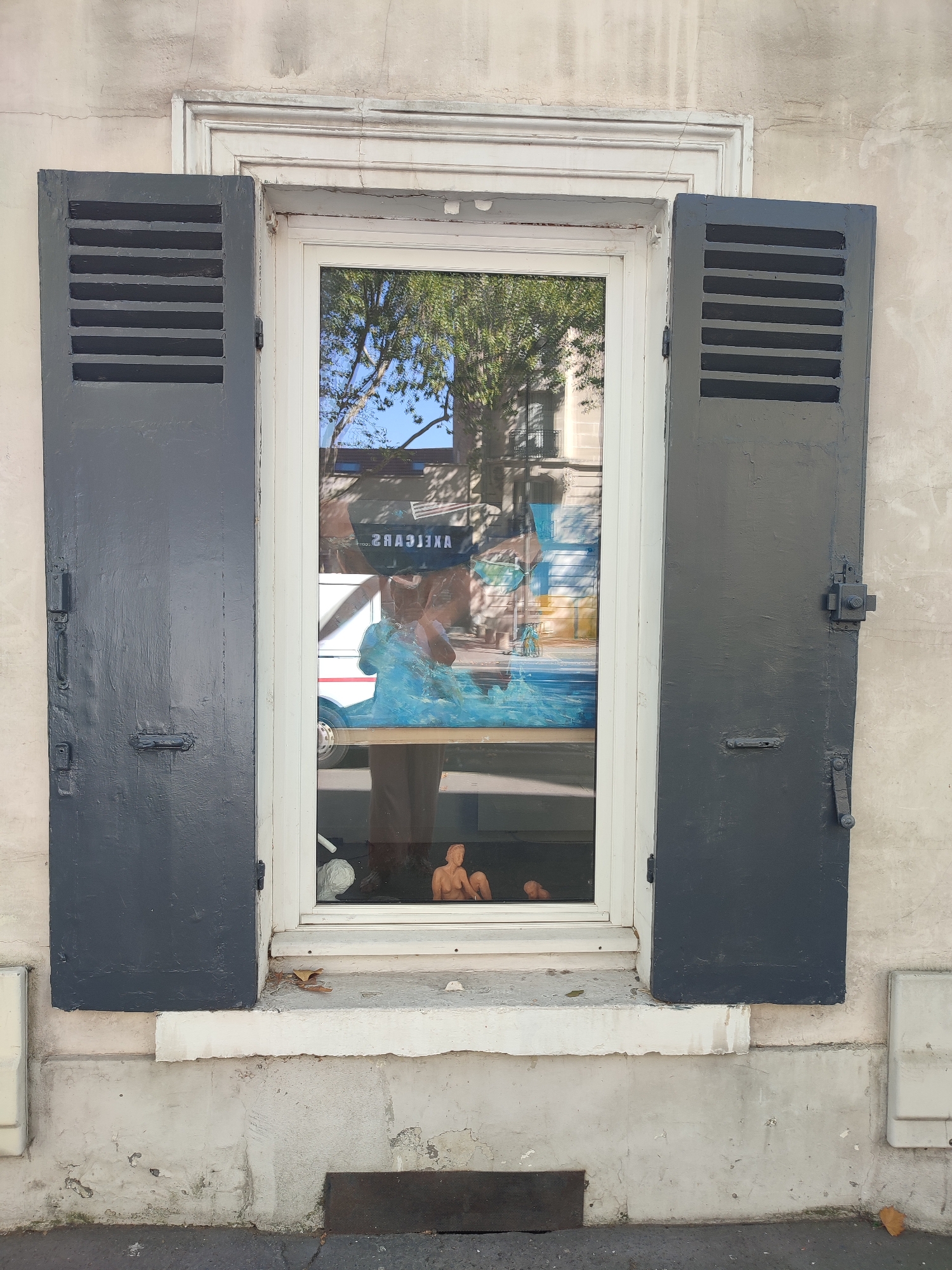 A medium shot, eye-level exterior view shows an open window with dark gray shutters on a weathered building facade in Boulogne-Billancourt, France. The window has a white frame and glass that reflects a tree and buildings across the street. Inside the window, a painting depicts a blue wave and yellow and white abstract shapes. In the foreground of the window, two small, nude figurines are visible, one of a woman and one of a child. A single brown leaf rests on the windowsill. The building facade is a light, textured color with cracks and peeling paint. The shutters are closed on the right and partially open on the left, revealing horizontal slats. The ground in front of the building is asphalt.