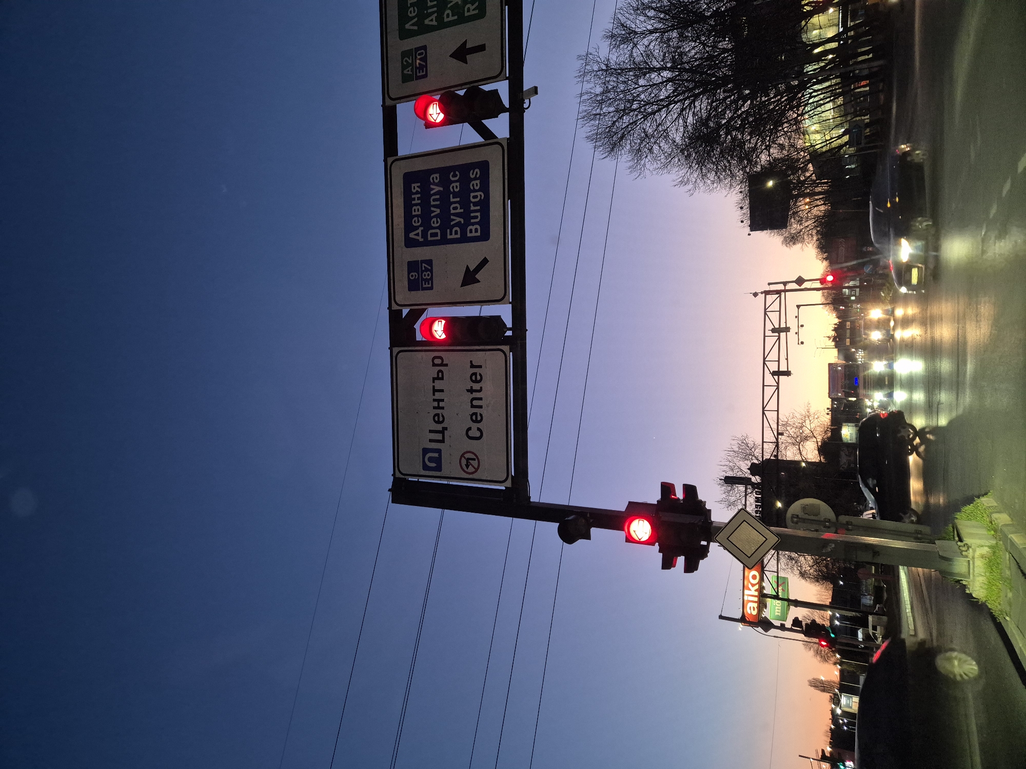 The image shows a traffic intersection in Varna, Bulgaria, at dusk or dawn. The sky is a gradient of blue and purple/pink. There are traffic lights showing red signals. Road signs indicate directions to "Center", "Devnya, Burgas", and "Airport (A2 E70)". A street sign with the word "aiko" is visible. Vehicles are waiting at the intersection.