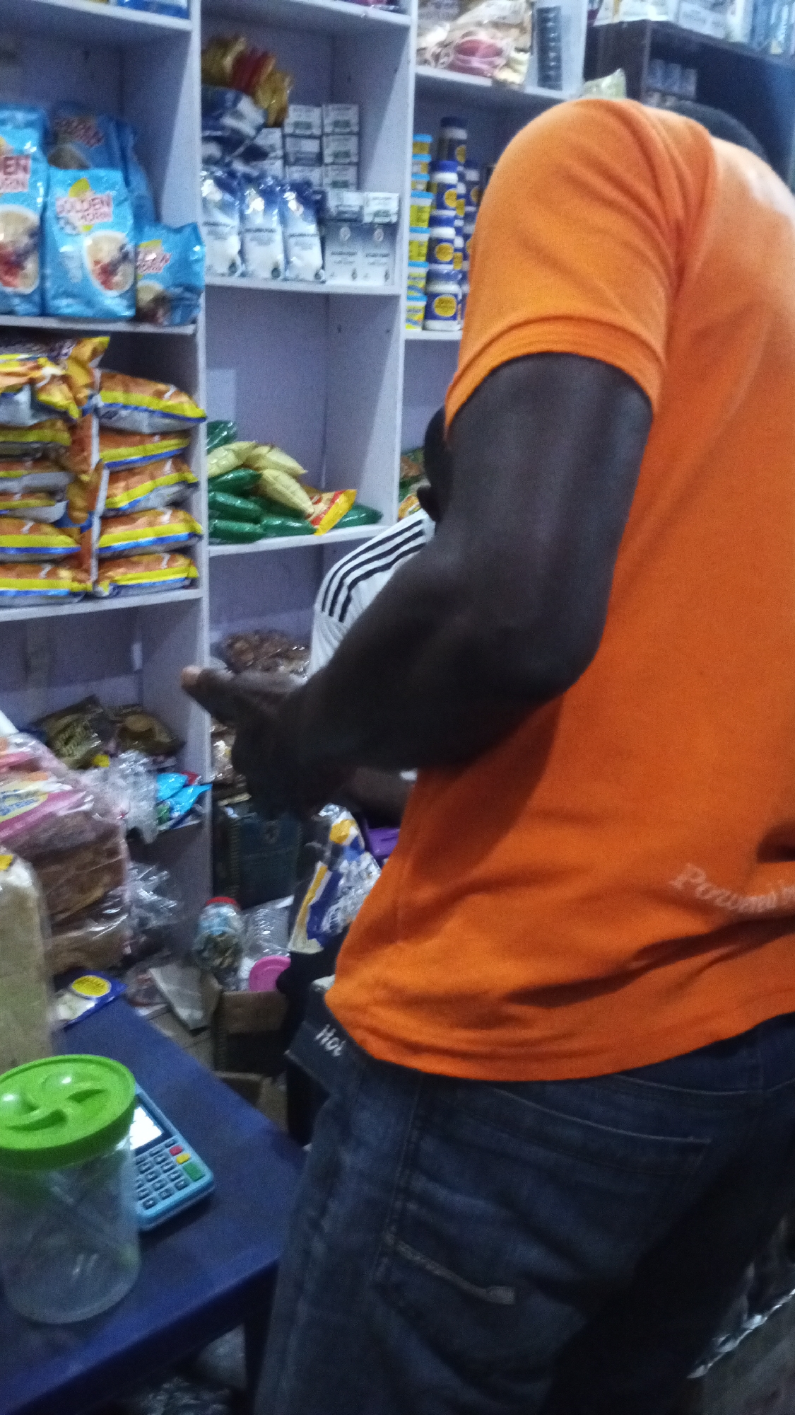A medium indoor shot in what appears to be a small general store in Adankolo, Nigeria, shows a person in an orange t-shirt and blue jeans in the right foreground, slightly obscuring the view. The person appears to be interacting with someone at a checkout counter to the left, indicated by the person's arm and hand gestures. The counter has a calculator and a clear plastic jar with a green lid. Behind the counter are shelves stocked with various food items including, but not limited to, cereal in blue boxes, stacks of bagged snacks, and yellow and green wrapped items. Another person is standing behind the person in the orange shirt.