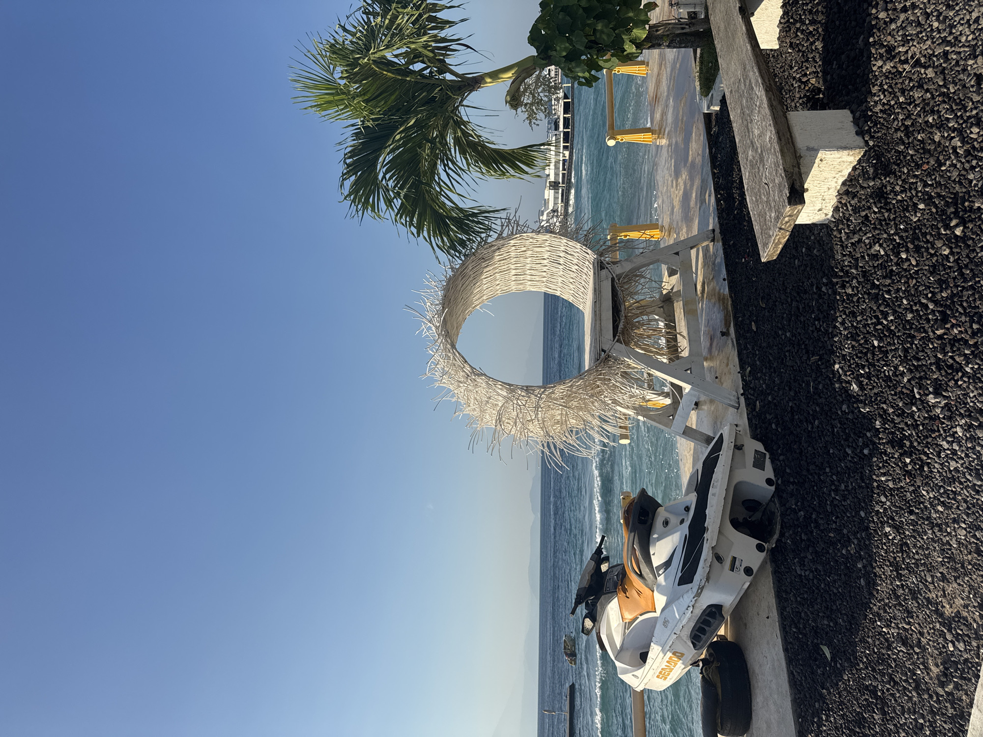 This image, captured in Nusa Penida, Indonesia, displays a coastal area under a clear, bright blue sky. The foreground consists of dark, small, uniform gravel. On the left, a white and orange Sea-Doo jet ski is positioned on a concrete platform, partially supported by a black tire. Adjacent to the jet ski, a large, white, circular, woven decorative structure, resembling a nest or wreath, stands on a white base on the same concrete platform. To the right of the platform, a green palm tree grows, and a rustic wooden bench with concrete supports is situated on the gravel. The concrete platform extends to meet the blue ocean, where small waves are visible, and yellow posts with ropes mark the edge. In the midground, the ocean extends to the horizon, with a large white ferry or ship present on the far right. Distant, hazy blue landmasses are visible across the water on the left side of the horizon. No individuals are present in the scene.