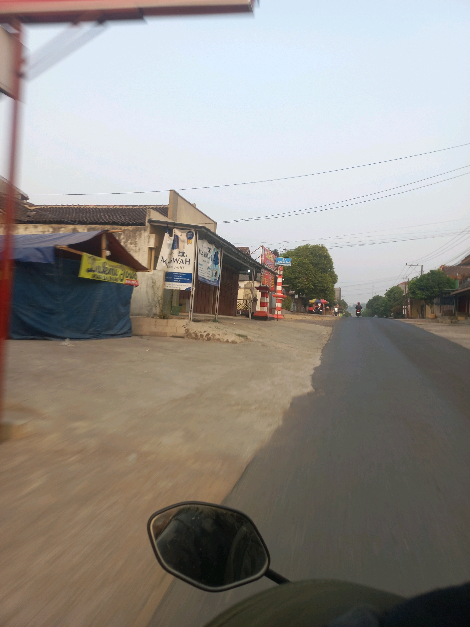 This is a street-level view, likely captured from a moving vehicle such as a motorcycle, in Ngarjosari, Indonesia. The image shows a paved road stretching into the distance, with a slight blur indicating motion. On the left side of the road, there are several buildings and stalls. One stall is covered with a blue tarp and has a yellow sign with text. Further down the street, there are signs for businesses, including one with "MARWAH JAHIT" visible. A tall, red and white striped pole stands near a building. Trees line the right side of the road, and more buildings are visible in the background. The sky is a light, hazy blue, suggesting it might be early morning or late afternoon. A single motorcyclist is visible in the distance on the road. A side mirror of the vehicle from which the photo is taken is visible in the lower foreground.