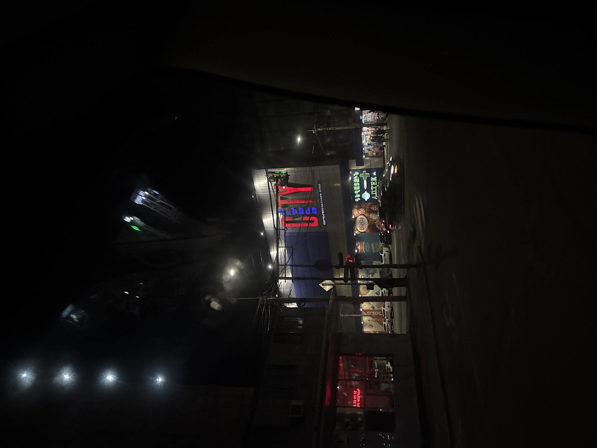 The image, taken at night, shows an urban scene in Yerevan, Armenia. Visible are buildings illuminated by streetlights and neon signs. A large sign with the word "CITY" in red is prominent, likely indicating a commercial establishment. Adjacent to it is another sign in Armenian script, possibly identifying a pharmacy ("Aptek"). Streetlights illuminate the roadway. The image appears to have been taken from inside a vehicle, looking out onto the street. Car headlights are visible in the bottom left. There are pedestrians on the sidewalk near the pharmacy.
