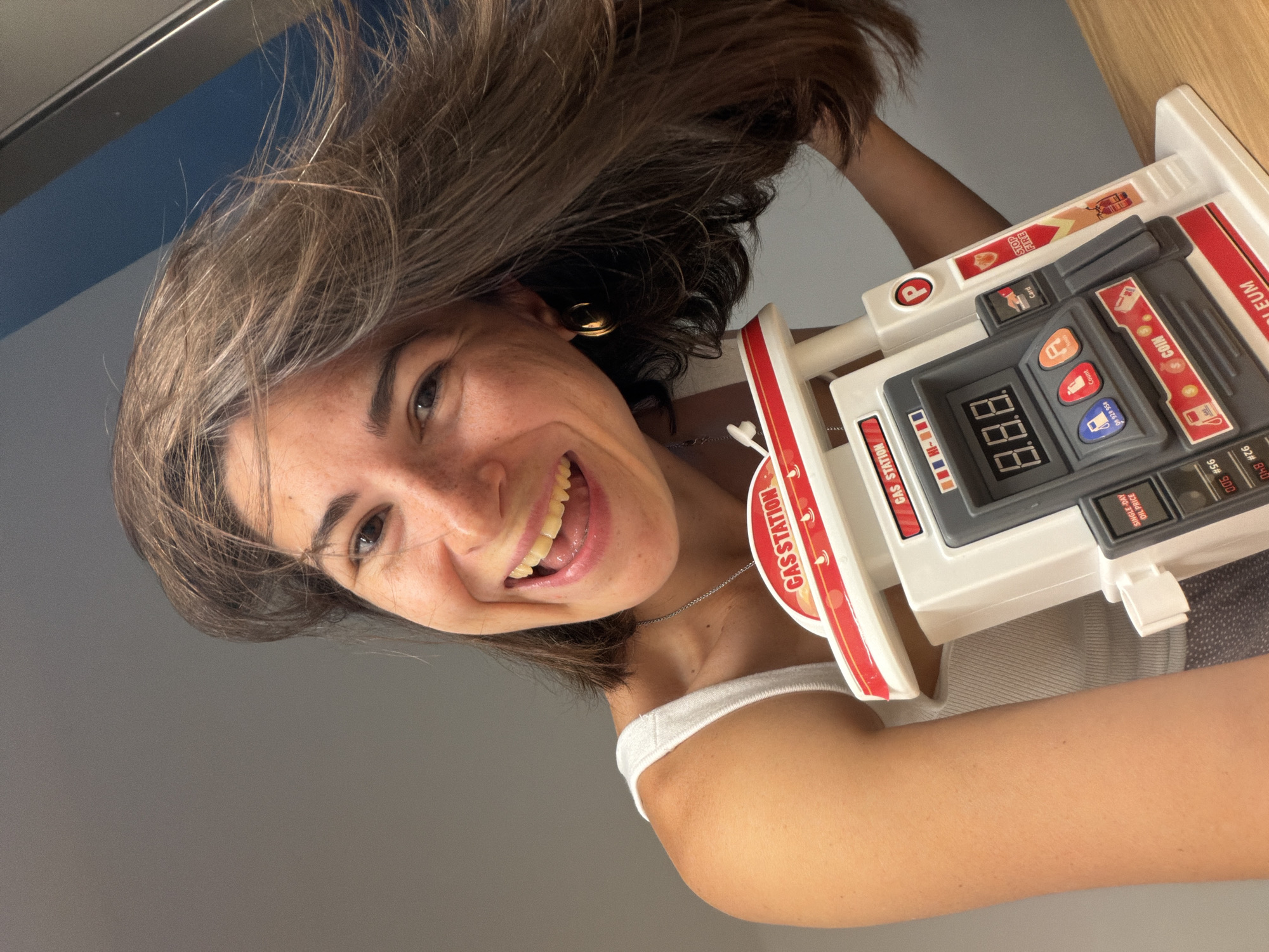 A smiling woman with brown hair, freckles, and a gold hoop earring is holding a toy gas station. The toy is white with red accents and includes buttons, a display showing "8.8.8", and stickers labeled "GAS STATION", "STOP FIRE", "COIN", and "SINGLE-DAY OIL PRICE". The background is a combination of blue and gray surfaces, likely walls. The woman is wearing a white tank top. The location is not provided in the image.
