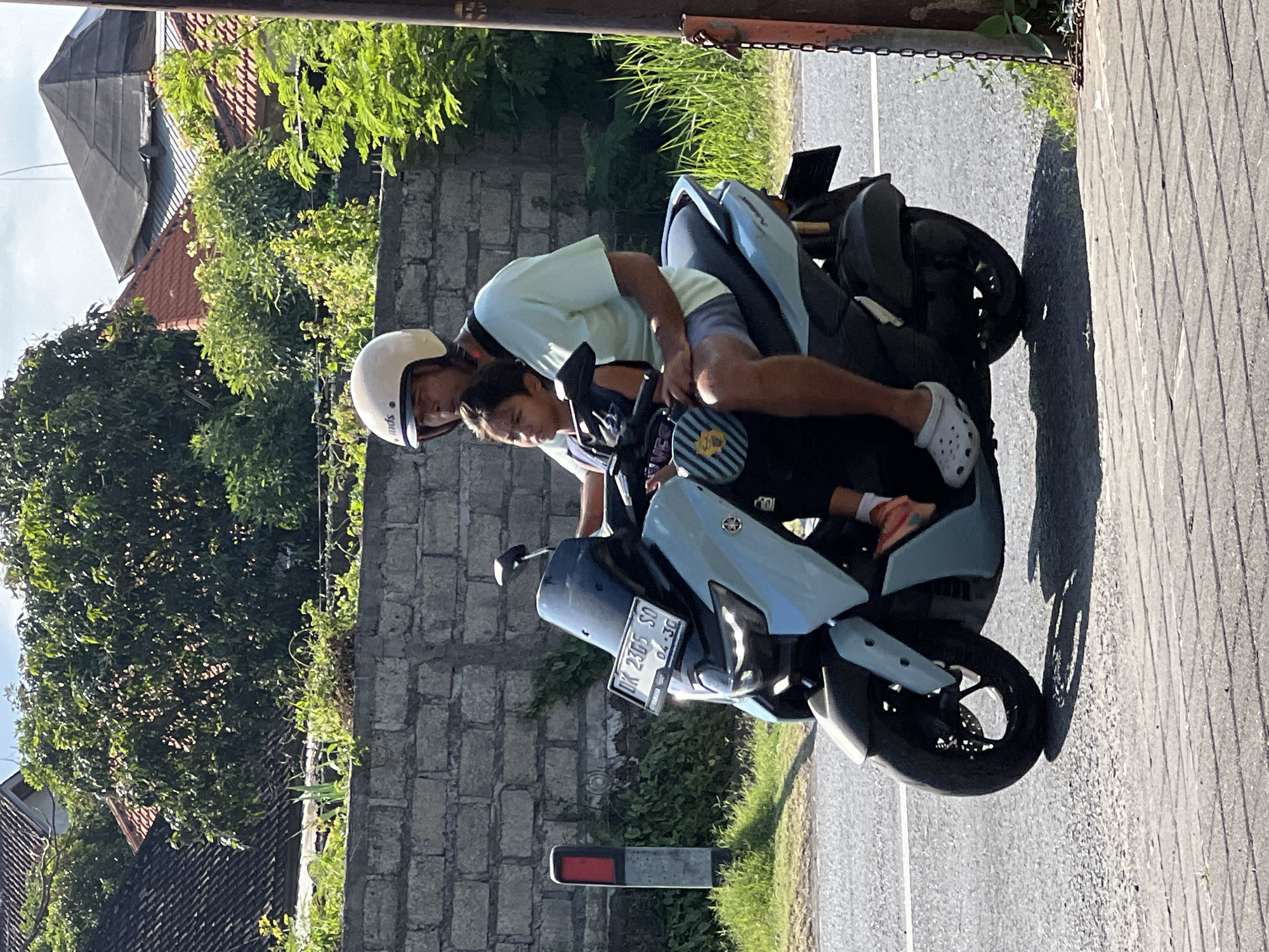 In Denpasar, Indonesia, a man wearing a light green t-shirt and grey shorts, along with a white helmet, is riding a light blue scooter. He is accompanied by a young boy who is seated in front of him, wearing a dark blue shirt. The scooter has a license plate that reads "DK 2365 SO". They are traveling on a road with a grey paved surface and a stone wall in the background. Green foliage and trees are visible behind the wall, and a few buildings with tiled roofs can be seen in the distance. The weather appears to be sunny, with a few clouds in the sky.