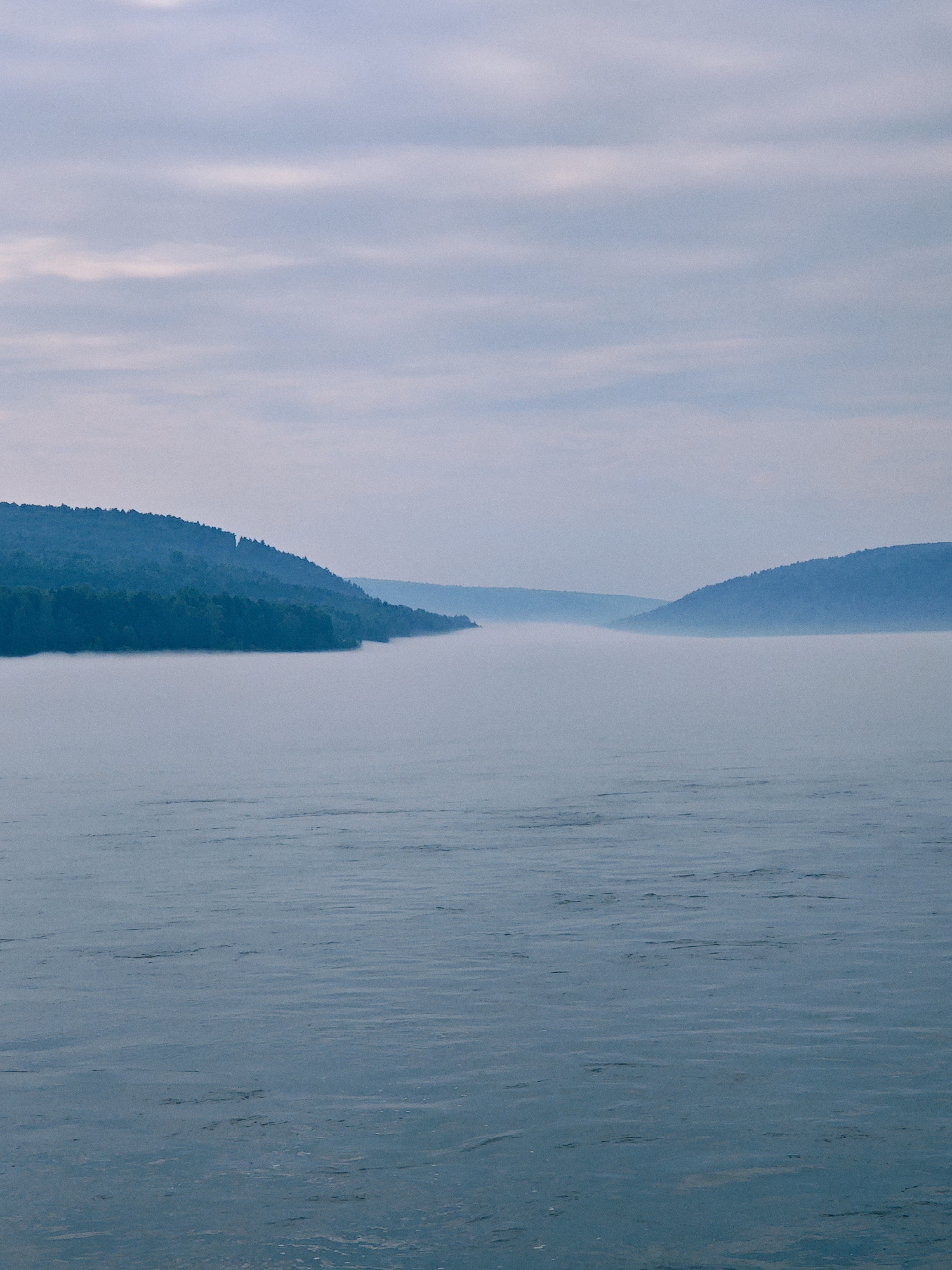 The image depicts a wide, calm body of water, likely a river or lake, under a cloudy sky. Rolling hills covered in dark green trees line the banks in the distance, fading into a misty haze. The foreground water has a textured surface with subtle ripples and reflections. The scene is dominated by cool blue and grey tones, suggesting an overcast day or early morning. The location is Atamanovo, Russia.