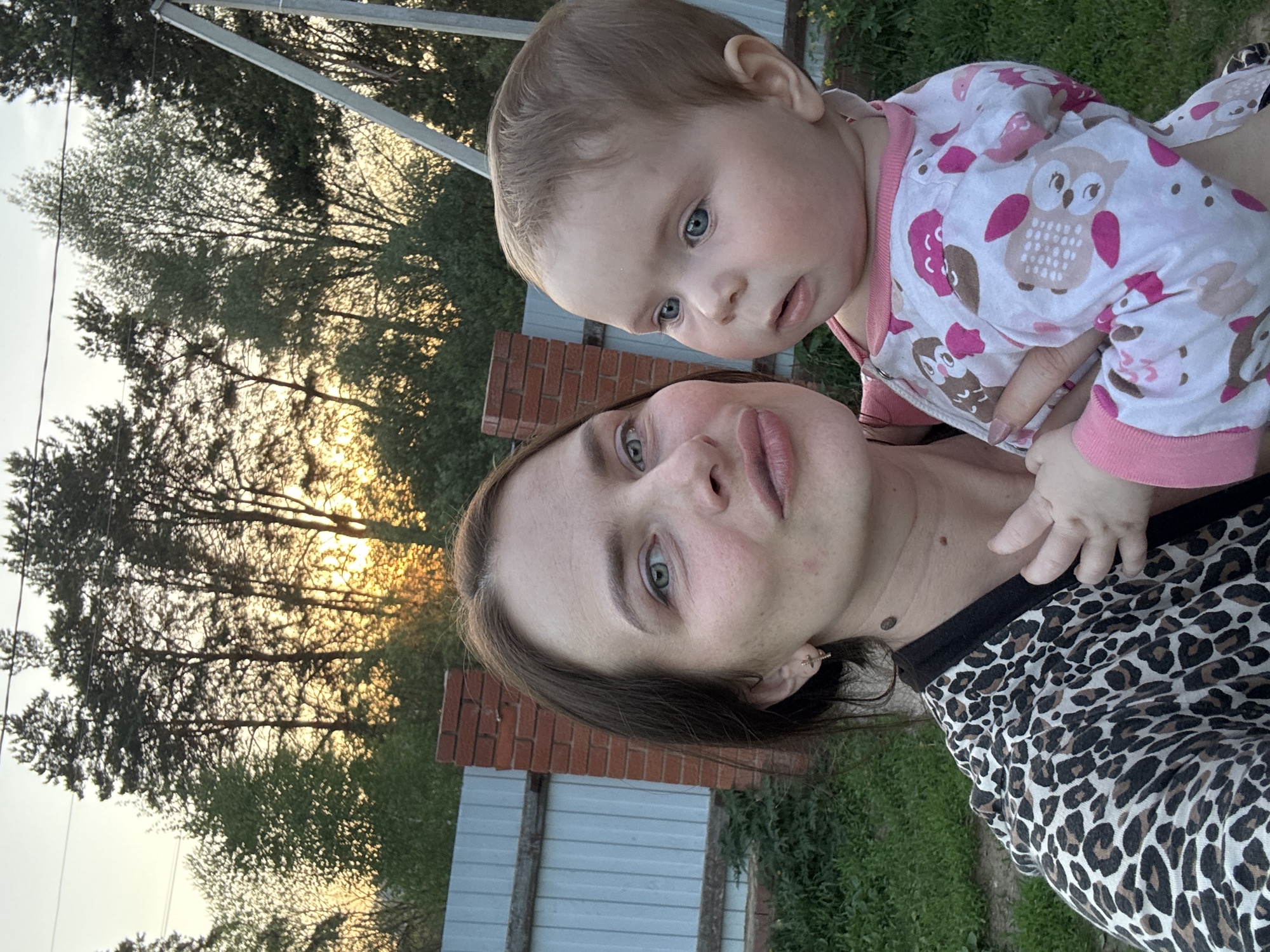 A woman is holding a baby in an outdoor setting, likely a yard or garden. The woman has brown hair and light eyes, and is wearing a leopard-print shirt. The baby has light hair and blue eyes, and is wearing pajamas with an owl pattern. Behind them, there is a fence made of brick and metal, and trees are visible in the background. The sky suggests it is sunset or sunrise.
The location is Rusalevka, Russia, though there are no elements to confirm it without additional information.