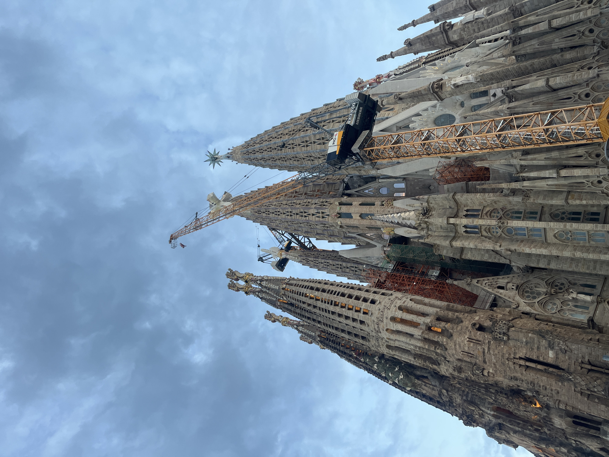 The image depicts a low-angle view of the Sagrada Familia basilica in Barcelona, Spain, under a cloudy sky. The basilica's iconic spires, characterized by their intricate Gothic and Art Nouveau architecture, dominate the frame. Several large construction cranes are visible, integrated with the structure, indicating ongoing construction or renovation work. One prominent yellow crane stands tall, while another black and yellow crane is positioned closer to the spires. Scaffolding and construction netting are also present on some parts of the building. The stone facade of the basilica is detailed, with numerous windows and sculpted elements. The lighting suggests it is either early morning or late afternoon. No people are clearly visible in the image.