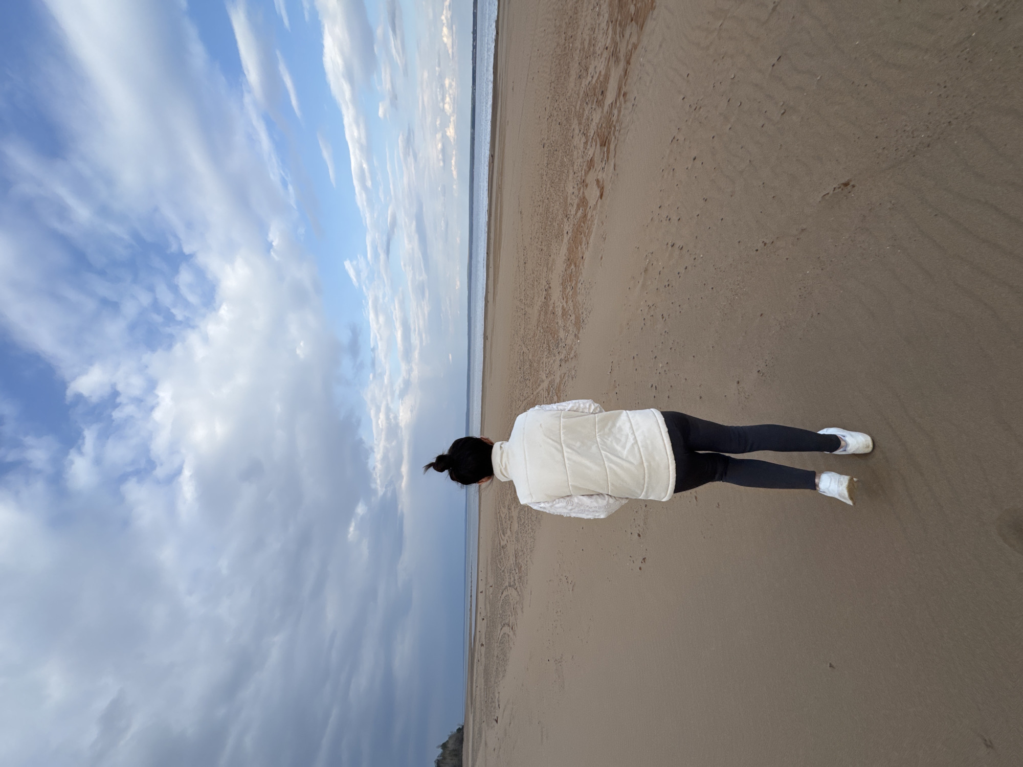 A woman stands on a sandy beach under a sky with scattered clouds. She wears a white vest over a sweater, black leggings, and white sneakers. Her hair is pulled back in a bun. The beach appears to stretch towards the horizon, with the sea visible in the distance. The location is near Noviy, Russia.