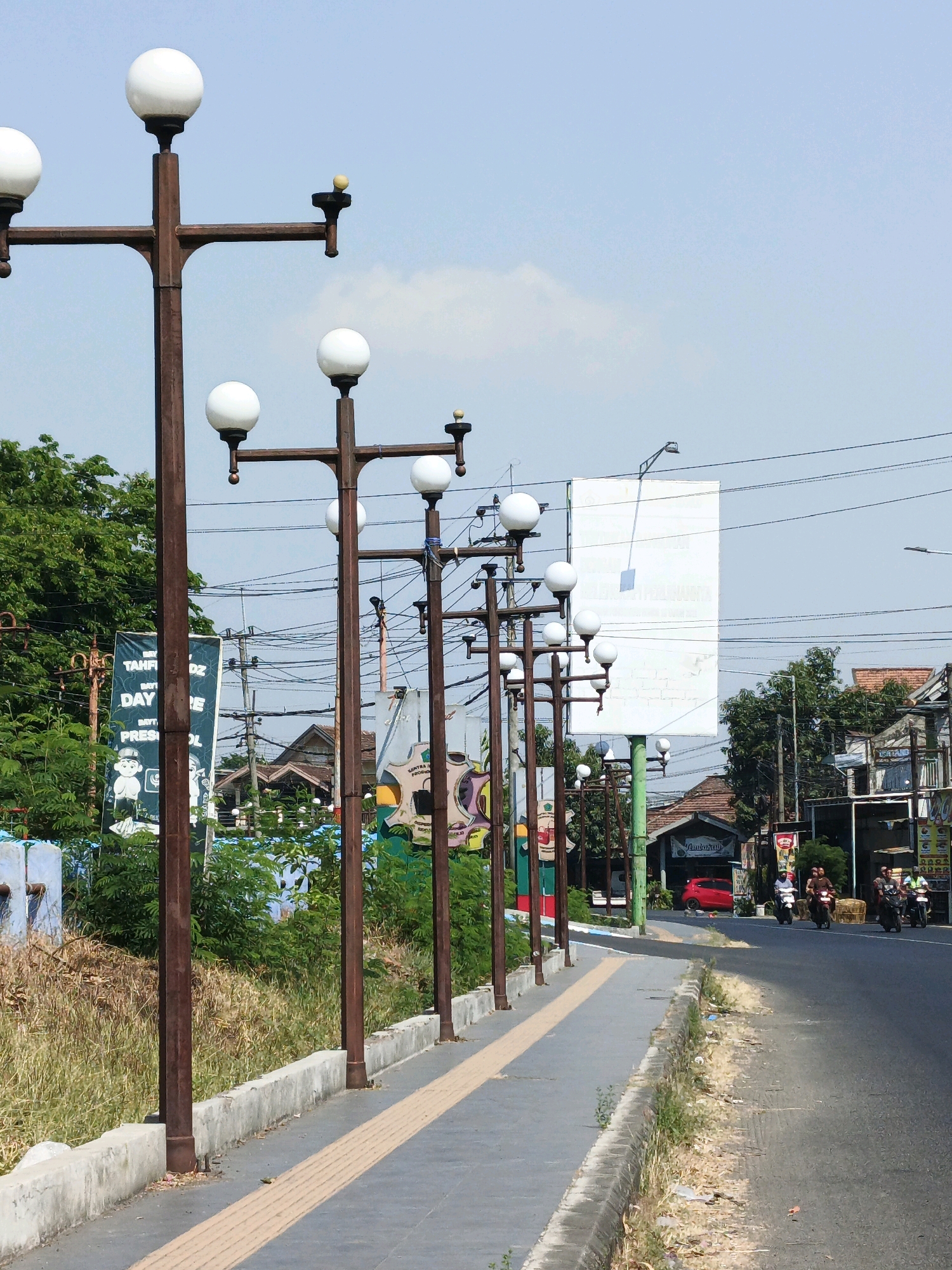 A low-angle, eye-level, medium shot captures a street scene in Randegan, Indonesia. The foreground is dominated by a sidewalk with a textured yellow strip, bordered by a concrete curb. Several weathered brown lampposts, each with multiple white spherical lights, line the sidewalk. Beyond the lampposts, a median with overgrown grass and some debris separates the sidewalk from the road.
On the left side of the frame, dense green foliage forms a backdrop, partially obscuring a tall, vertical banner with text in Indonesian and cartoonish figures. Further back, a variety of buildings with tiled roofs are visible, along with a prominent, large white billboard. A network of utility poles and wires crisscrosses the sky.
The road on the right side of the image features several motorcycles with riders. The weather appears to be clear and sunny, indicated by the bright light and the presence of shadows. The overall impression is of a typical street scene in a developed area of Indonesia.
