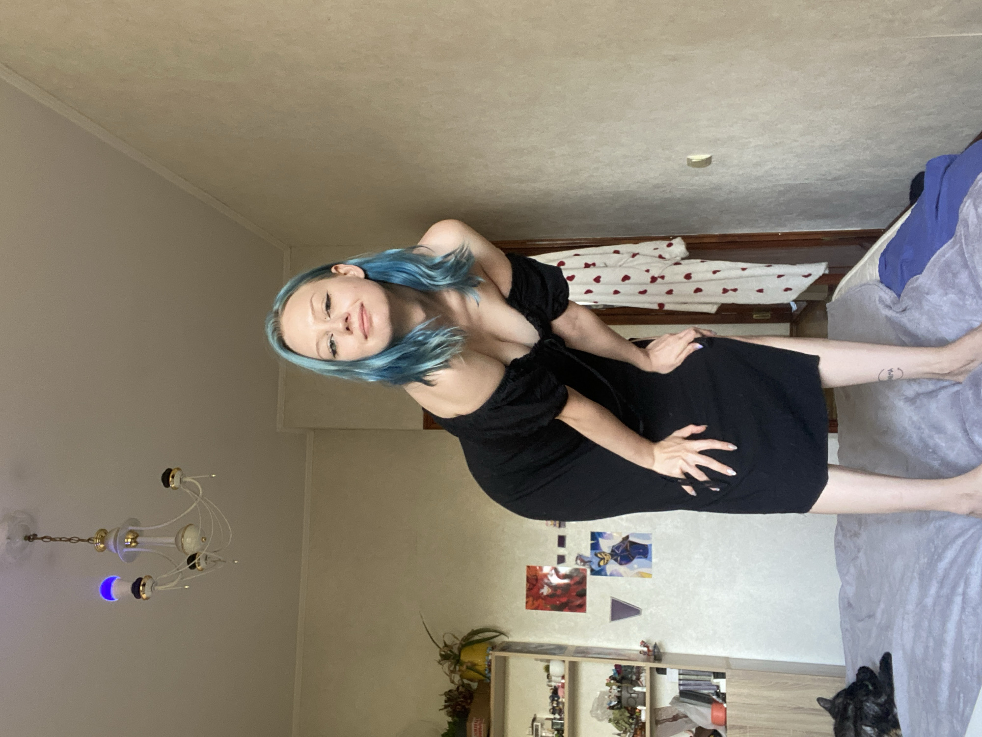 A woman with blue hair, wearing a black dress, is lying on a bed. She is leaning on her arm and looking at the camera with a slight smile. The room appears to be a bedroom, with a bed, a dresser with shelves filled with items, and a chandelier on the ceiling. There are also some framed pictures on the wall. A cat is visible lying on the bed. The lighting suggests it is daytime. The overall mood appears relaxed.