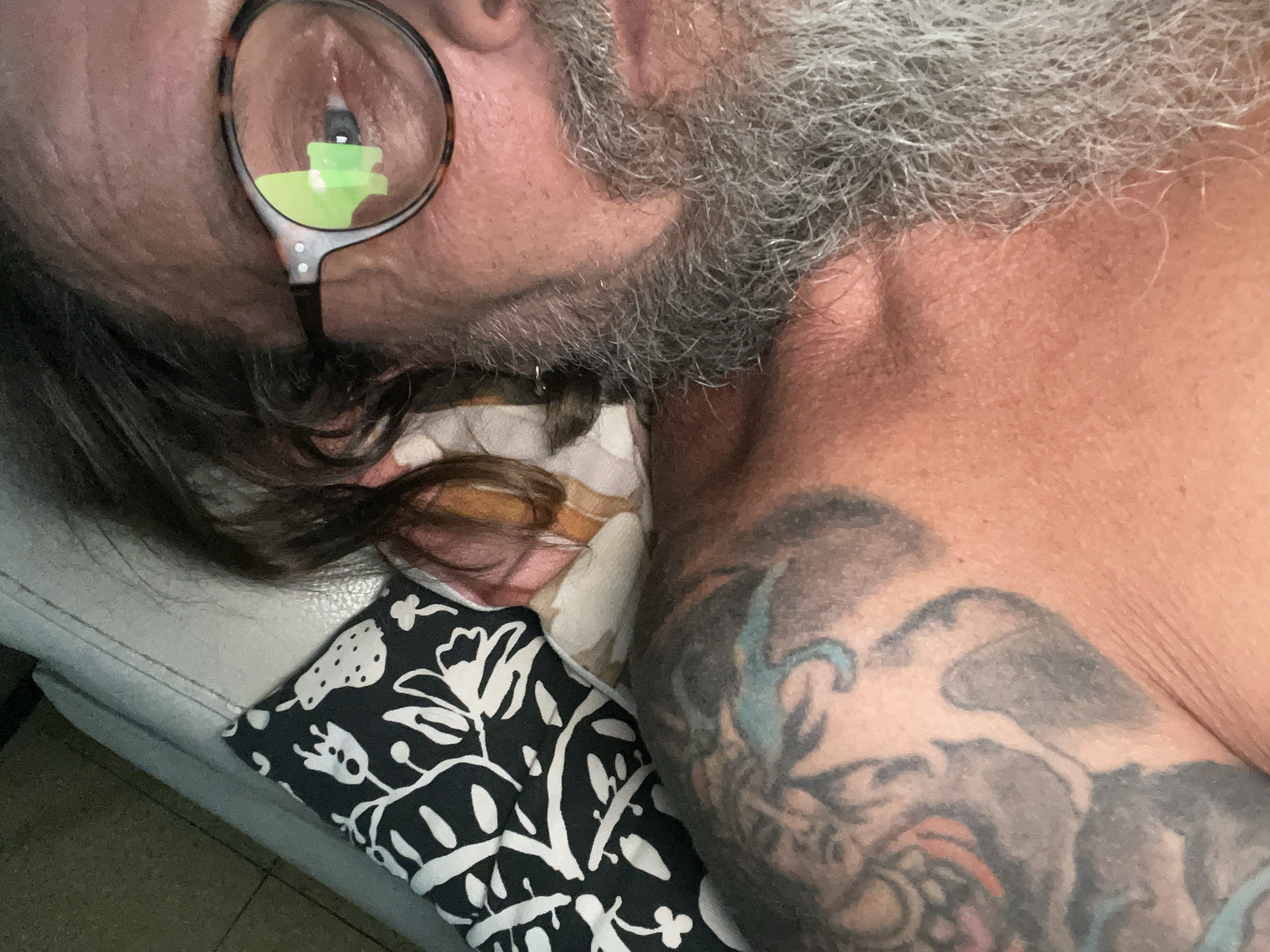 A close-up, selfie-style image shows a person from the chest up, with their head tilted towards the camera. They are wearing glasses with tortoise-shell frames, which reflect a bright green object. The person has a grey, full beard and long, dark brown hair that partially covers their face. The skin appears flushed and sun-kissed, with visible texture. On the left shoulder and upper chest, there is a large, colorful tattoo depicting what appears to be a dragon or serpent. The person is lying down, with a patterned pillow and a white cushioned surface visible in the background. The pillow has a black and white floral design. The overall impression is casual and intimate. The image was taken in Netanya, Israel.