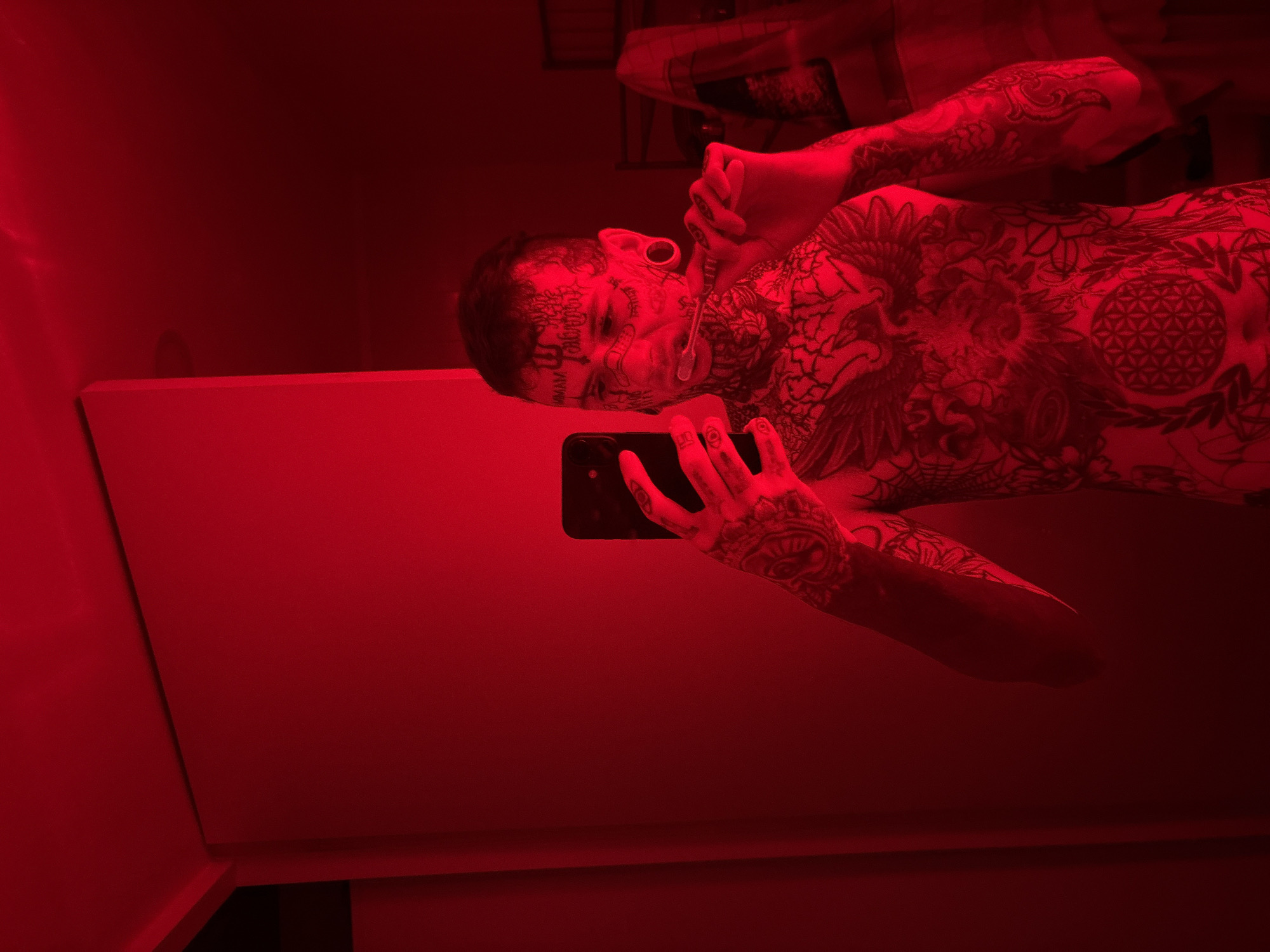 A shirtless person with extensive tattoos, including facial tattoos, is holding a smartphone in their right hand, seemingly taking a selfie. They are positioned in what appears to be a bathroom, with a red light illuminating the scene. The location of the image within Buenos Aires, Argentina, is not identifiable from the given visual information.