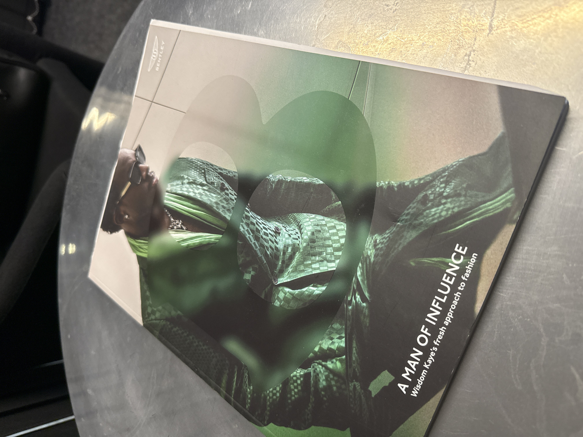 A magazine or booklet with a cover featuring a person dressed in green checkered patterned clothing is placed on a gray surface. The cover also features a green shaded design and the text "A MAN OF INFLUENCE" with the caption "Wisdom Kaye's fresh approach to fashion". The Bentley logo is visible in the top left corner of the cover. The image is taken indoors, with a black object visible on the left side. The location is unknown, and there is no information to suggest the location is in Plovdiv, Bulgaria.