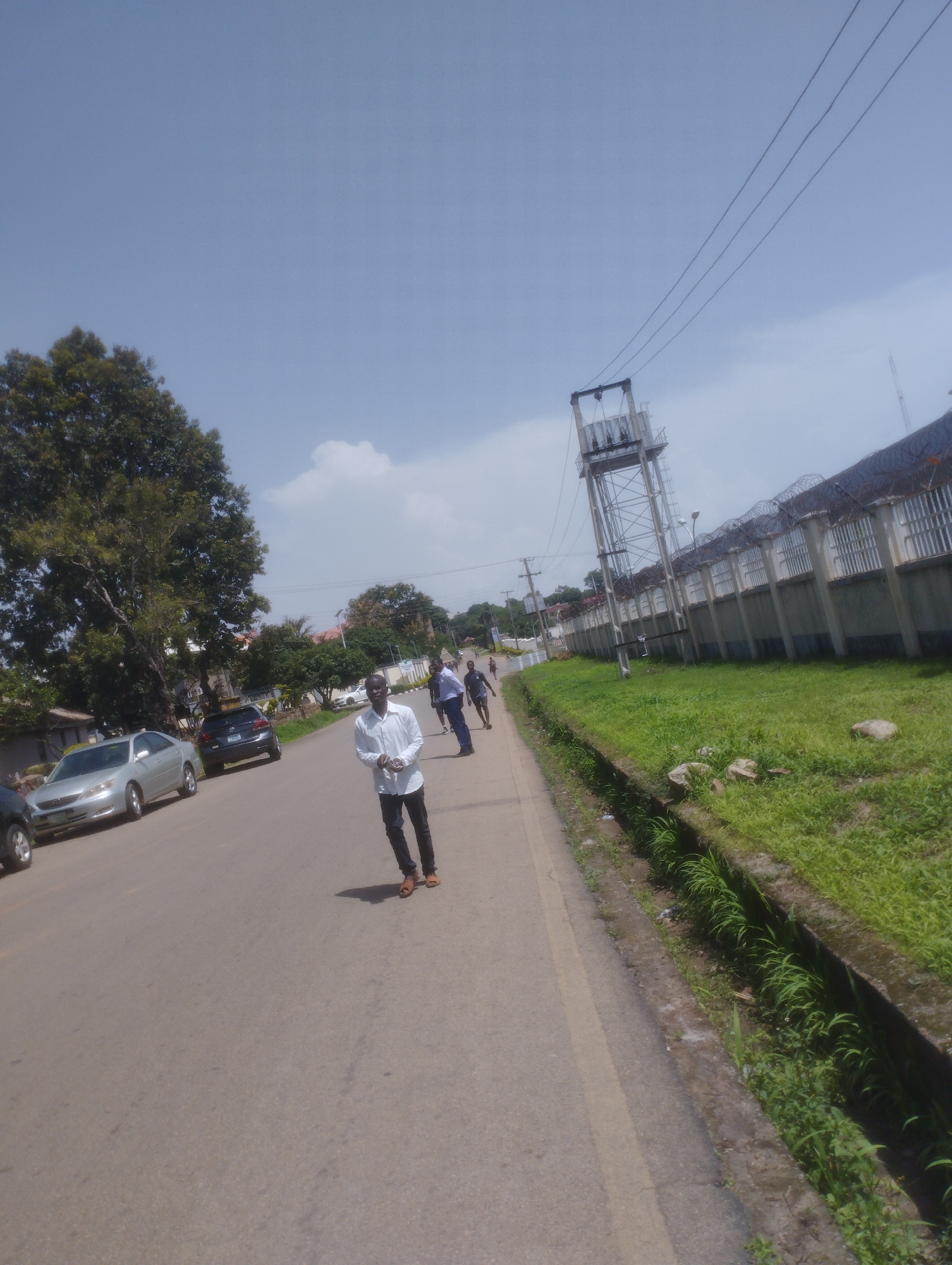 The image depicts a street scene in Jos, Nigeria, on a bright, clear day. In the foreground, a man in a white shirt and dark pants walks along the asphalt road, facing the camera with a neutral expression. Several cars are parked on the side of the road, including a silver sedan and a dark SUV. To the right of the road, there is a grassy area with a concrete drainage channel. Beyond the grass, a long, fortified wall with barbed wire on top extends into the distance, with a utility tower visible behind it. The sky is blue with a few scattered clouds. In the background, trees and buildings suggest a suburban or urban environment. Another utility pole with electrical wires is visible on the left side of the road.
