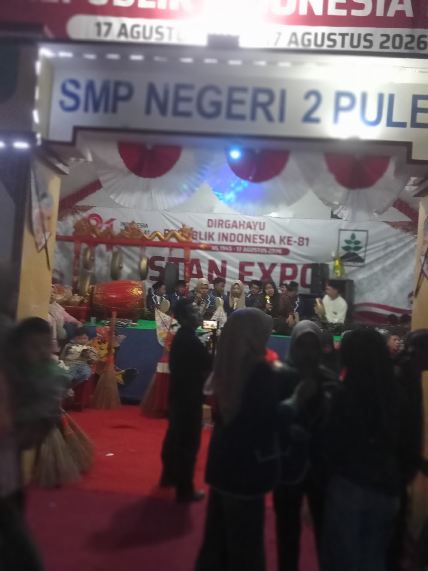 This is a blurry, medium-distance, eye-level shot of an outdoor event, possibly a school fair or exhibition, in Pule, Indonesia. The foreground is dominated by a bright red carpet, with several people in dark clothing standing on it, facing away from the camera. To the left, a group of people, including children, are seated on a raised platform, some of them holding brooms. Behind them, a traditional Indonesian gamelan musical instrument, likely a gong, is visible.
Further back, a large banner is displayed with text that appears to be in Indonesian, featuring "DIRGAHAYU REPUBLIK INDONESIA KE-81" which translates to "81st Anniversary of the Republic of Indonesia". Below this, the words "STAN EXPO" are prominently displayed. Above the banner, a sign reads "SMP NEGERI 2 PULE," indicating the location as Pule State Junior High School 2. The overall atmosphere suggests a festive or celebratory occasion, with elements of Indonesian culture and education evident.