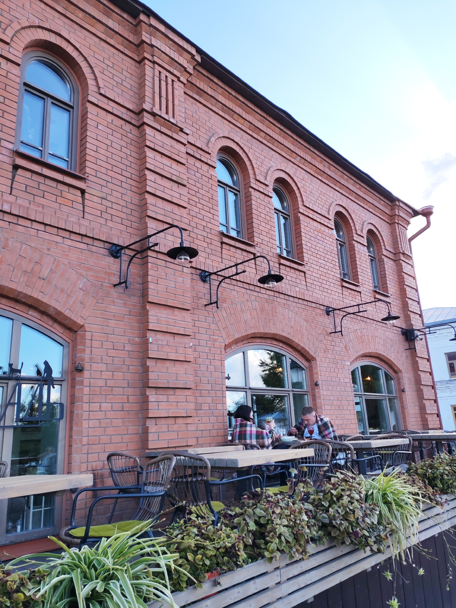 In the city of Plyos, Russia, a low-angle, full-shot photograph captures the exterior of a two-story brick building with an outdoor cafe seating area in the foreground. The building, constructed of reddish-orange brick, features arched windows on both levels. Several wrought-iron outdoor light fixtures are mounted on the brick facade above the lower windows. Below these, a railing separates the cafe from the street.
The cafe area is furnished with wooden tables and chairs. In the center of the frame, a man and a woman are seated at one of the tables, facing each other. The woman, on the left, is wearing a plaid blanket over her shoulders. The man, on the right, is wearing a plaid shirt and is looking down at the table. They appear to be eating or drinking. The tables and chairs are arranged along the front of the building, with a wooden planter box filled with greenery running along the edge of the cafe area. The sky above is a clear, pale blue.