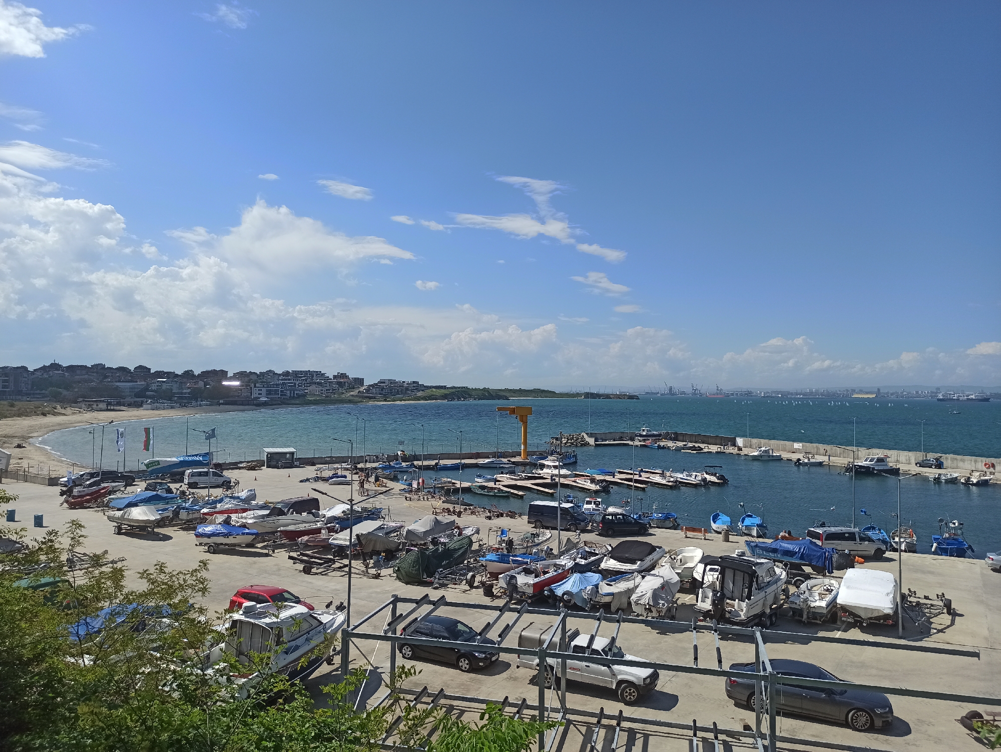 The image shows a harbor area in Burgas, Bulgaria on a clear, sunny day. In the foreground, numerous boats are parked on trailers, along with several cars. A concrete pier leads into the harbor, lined with docked boats and a few people walking. In the distance, the Burgas coastline is visible, featuring buildings and industrial port infrastructure. The sky is blue with scattered clouds.