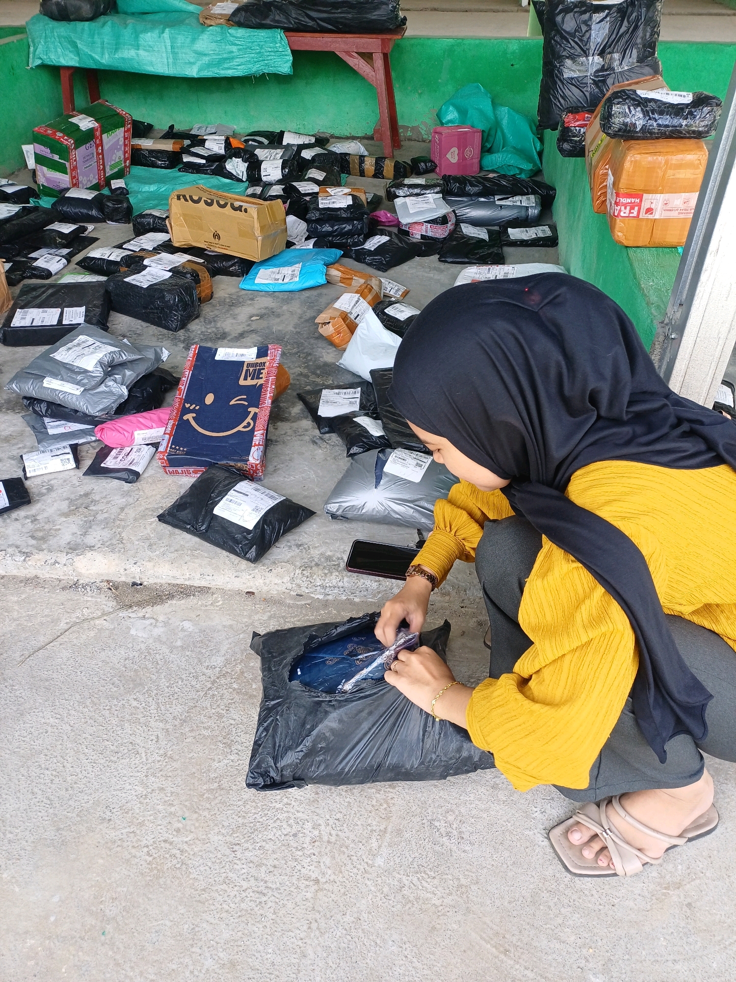 A woman wearing a black hijab and a yellow shirt is crouching on a concrete floor surrounded by numerous packages. She is in the process of opening one of the packages, which is made of black plastic. The packages vary in size and shape, with some appearing to be wrapped in bubble wrap and others in plastic mailers. Several are labeled with shipping information. In the background, there is a green wall and a wooden structure. The scene appears to be an outdoor or semi-outdoor area, possibly a delivery hub or storage space. The overall impression is one of a busy shipping or sorting operation. The location is Pasir Belengkong, Indonesia.
